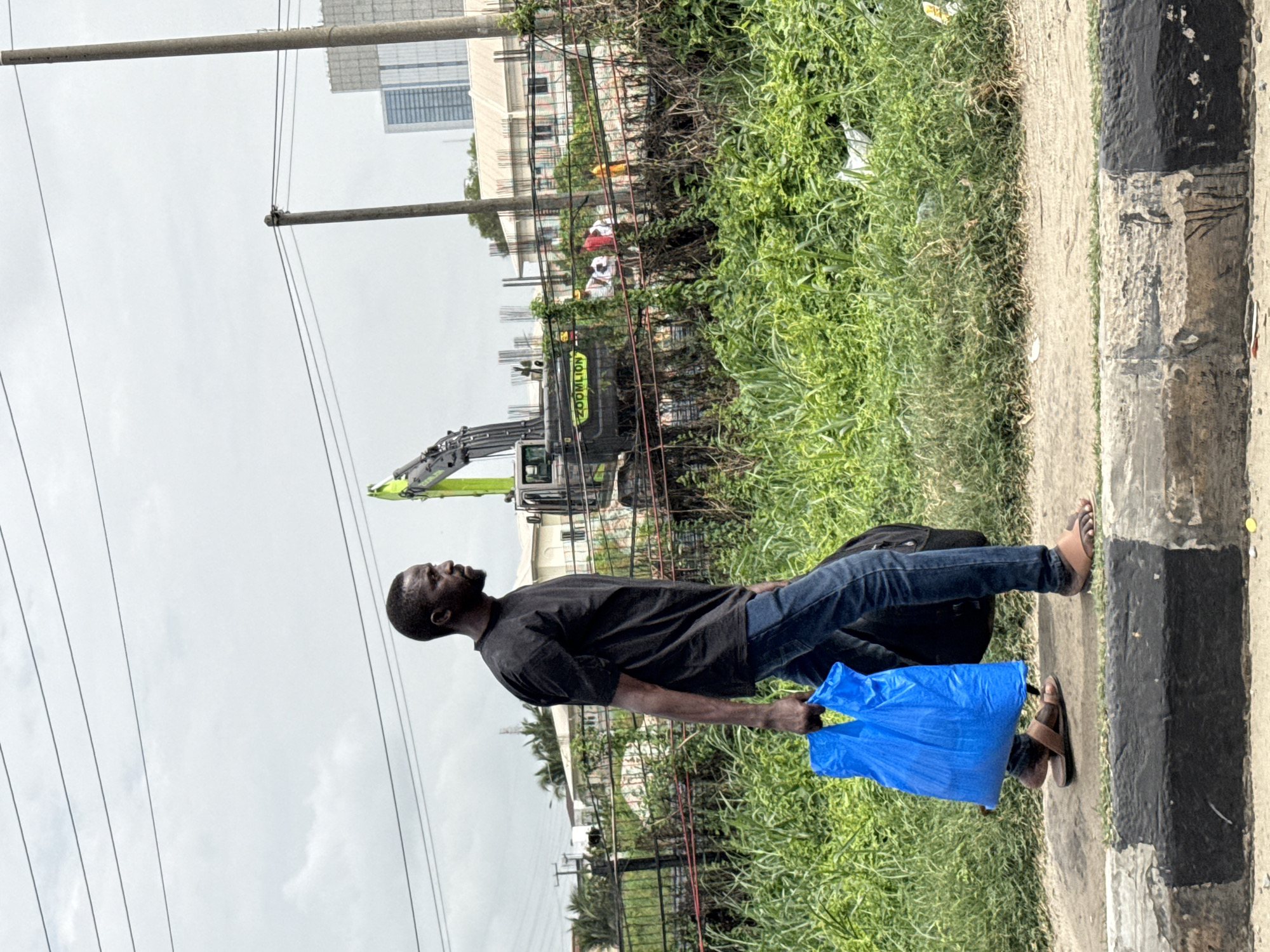 A man is walking along a sidewalk in Ilado, Nigeria. He is wearing a black t-shirt and blue jeans, and he is carrying a blue plastic bag in his left hand and a black backpack on his back. He is walking near a construction site, where a large excavator with the word "ZOOMLION" on it is visible. There is dense green vegetation along the side of the sidewalk. Overhead, power lines stretch across the sky. In the background, buildings can be seen.