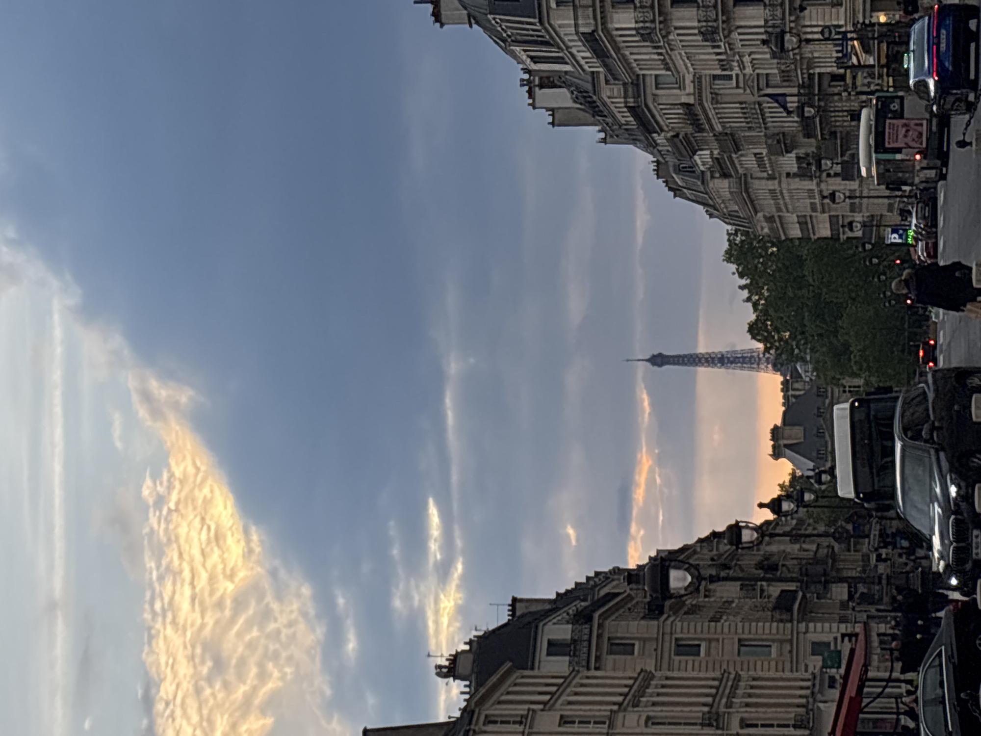 The image is taken in Paris, France, featuring the Eiffel Tower in the distance, framed by buildings on either side. The sky is predominantly blue, with streaks of white and orange clouds. The buildings are multi-storied and of traditional Parisian architecture. Vehicles are parked along the street. A person walks on the sidewalk.