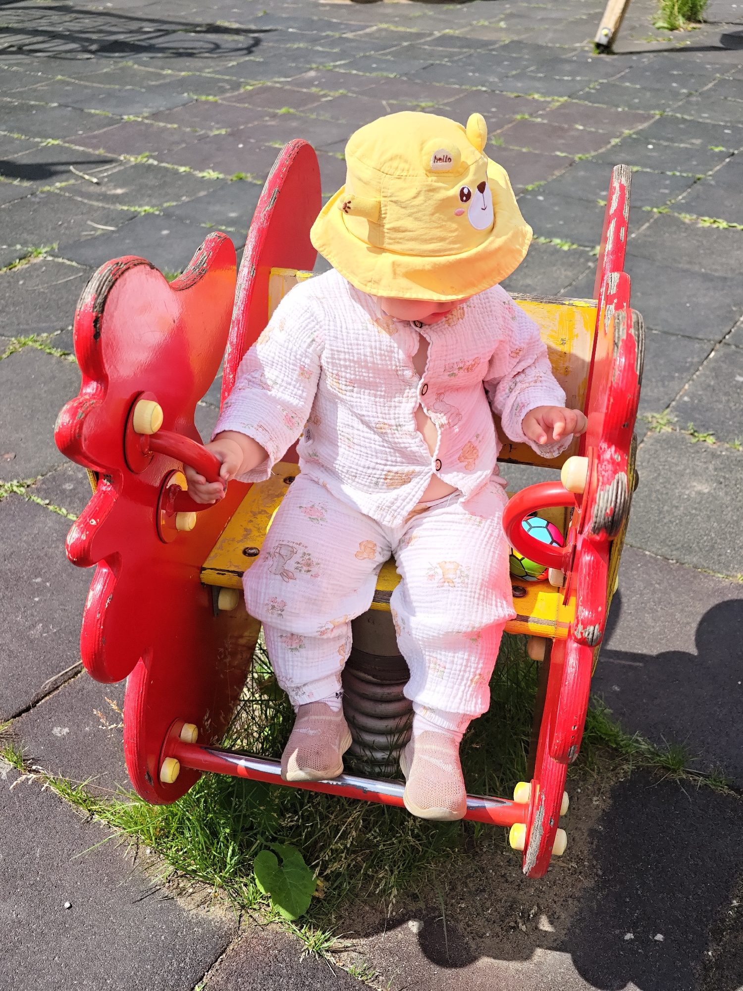 A baby wearing a yellow sun hat with a bear design and a white outfit with a floral pattern sits on a red and yellow spring rider on a playground. The playground surface is paved with dark grey interlocking rubber tiles and some patches of grass. The baby is holding onto a red handle on the side of the rider. The spring rider is shaped like a mythical creature with large red wings. The scene is outdoors on a sunny day.