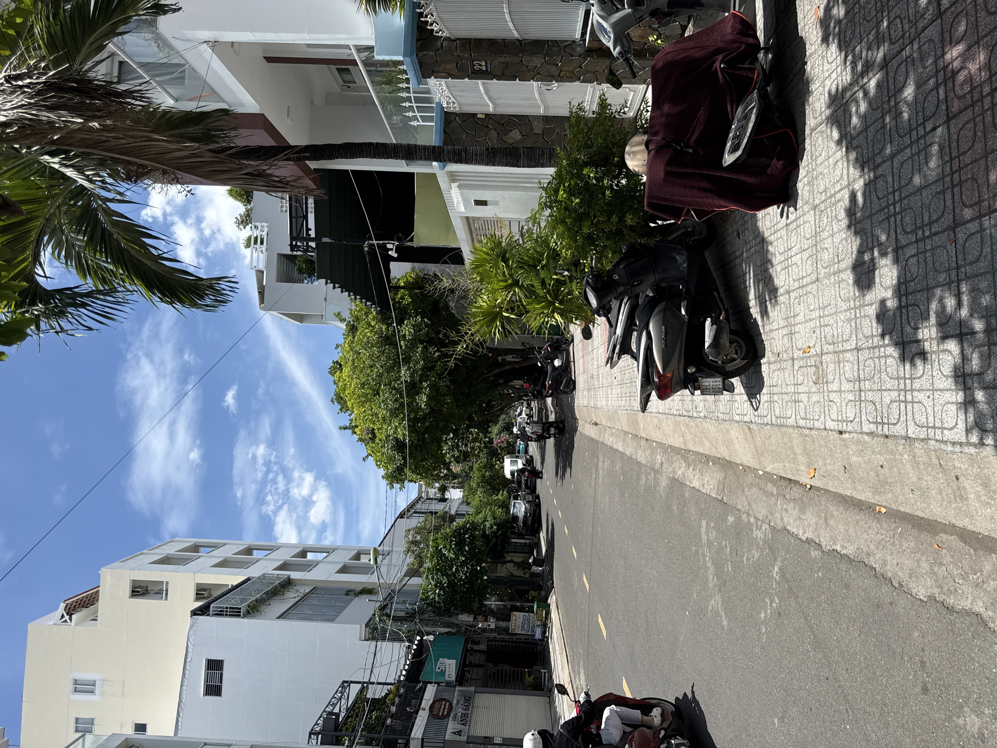 This is a medium-shot, street-level, eye-level, horizontal view of a street in Nha Trang, Vietnam. The scene depicts a residential and commercial area under a bright, blue, partly cloudy sky.
On the left side of the street, a row of multi-story buildings lines the road. The nearest building is white with multiple windows and balconies. Further down the street, a taller, cream-colored building is visible. The street is paved with asphalt, marked with yellow lines. A few motorcycles are parked along the sidewalk, and one person on a red motorcycle is visible on the left.
The right side of the street features more buildings, including a modern white house with a stone fence and a gate numbered "22". Lush green trees and bushes are abundant, with a prominent palm tree in the foreground. Several parked motorcycles are on the sidewalk, some covered with blankets. The sidewalk is paved with square tiles.
The overall impression is a typical urban street scene in Vietnam on a sunny day, characterized by architecture, transportation, and vegetation.