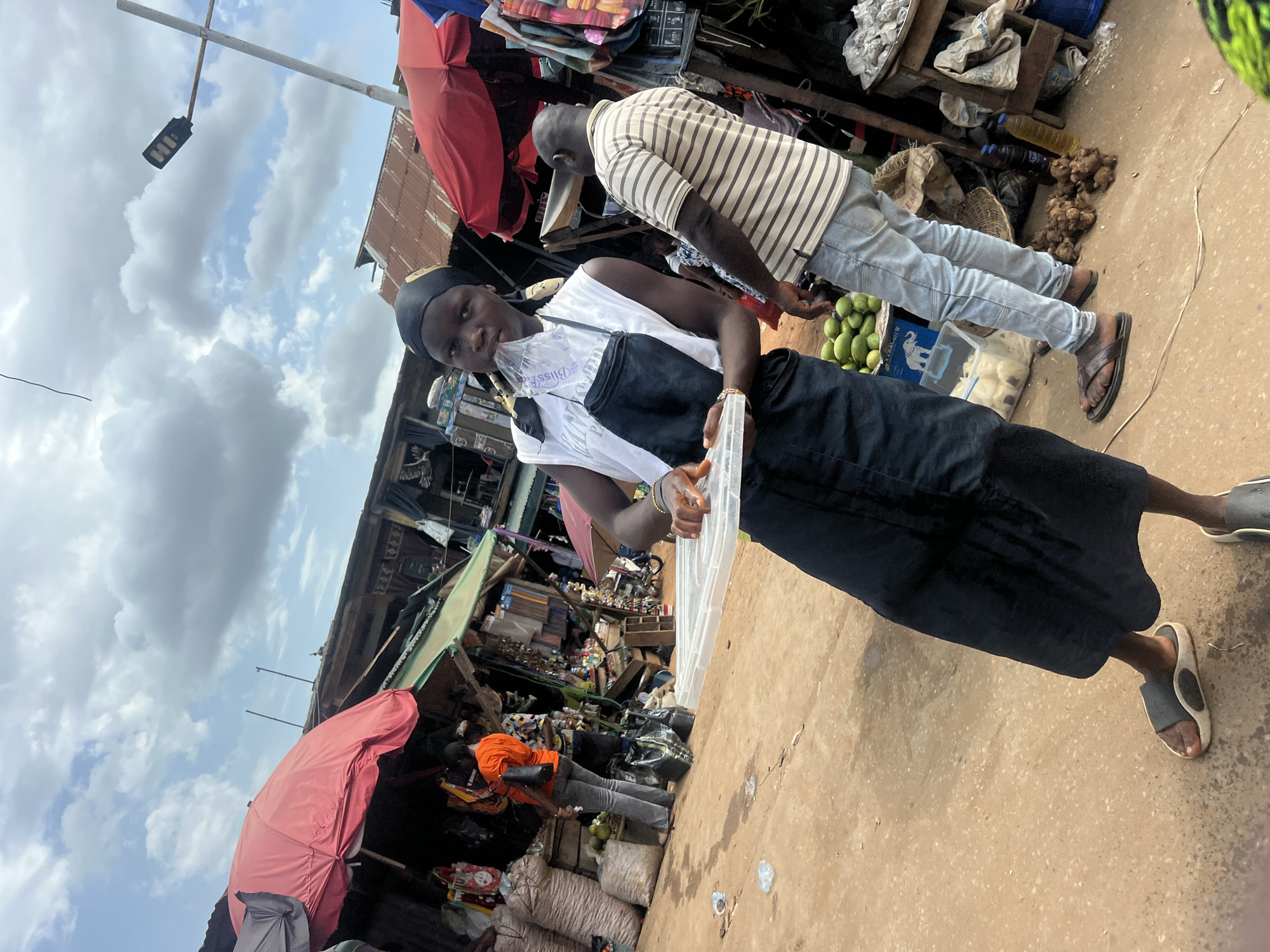 A medium shot shows a young woman holding clear plastic containers, centered and facing forward in an outdoor marketplace. She wears a white tank top, a black dress, a black head covering, and sandals. A man stands behind her to the right, wearing a striped shirt and light blue jeans. In the background are various market stalls with goods on display, including red umbrellas, assorted merchandise, and produce. The ground appears to be a dusty or sandy surface. The sky above is partly cloudy.
