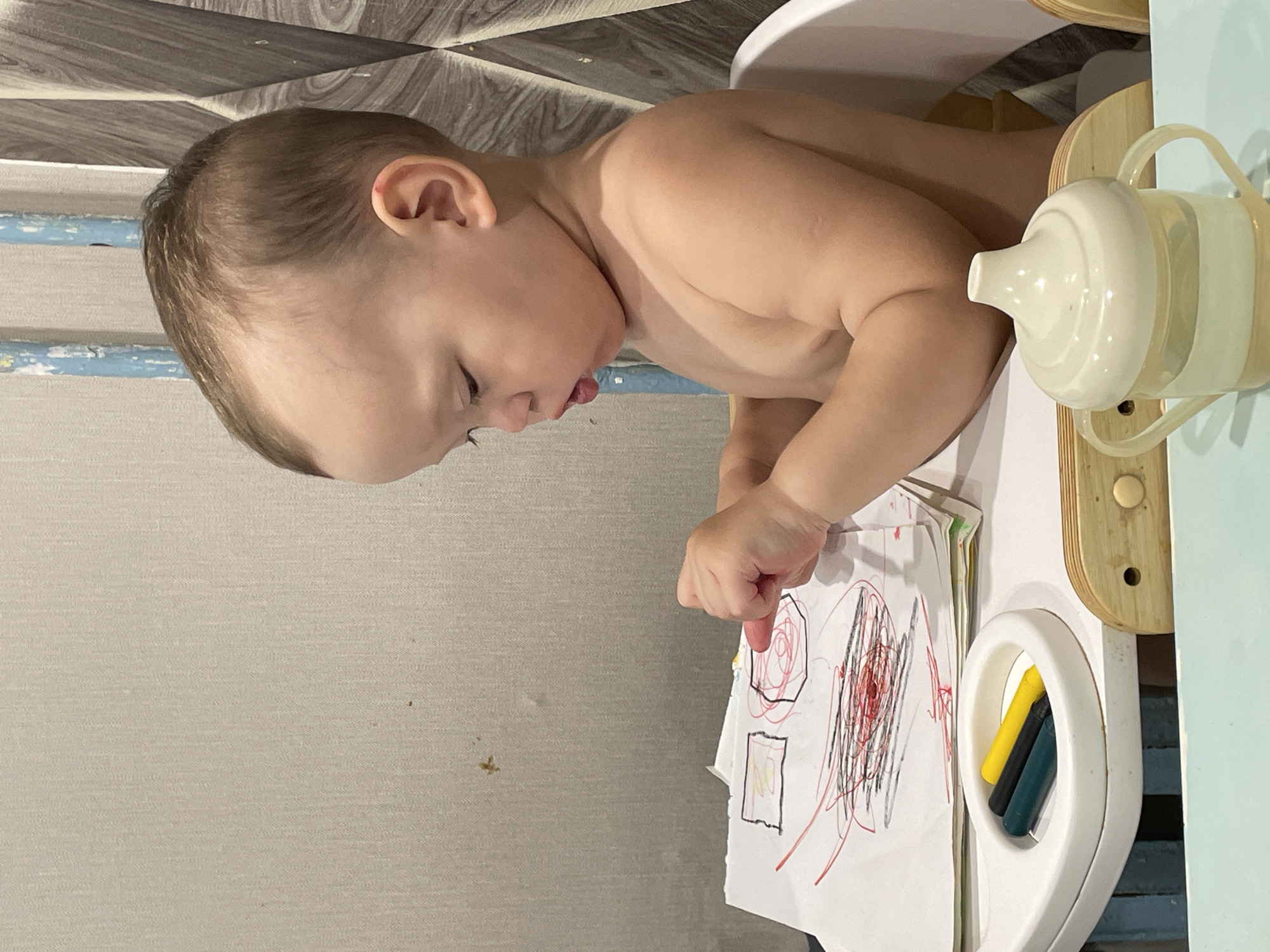 A fair-skinned, naked toddler sits in a white high chair at a small table. The toddler's head is turned to the left as the toddler looks at a white piece of paper with red, black, and grey scribbles. The index finger of the toddler's right hand points at the paper. A white sippy cup sits at the right side of the table. A tray attached to the high chair holds yellow and black crayons. The background includes a grey wall with a textured marble-like surface, and a blue wall with a grey texture. The location is likely a private residence in Perm, Russia.