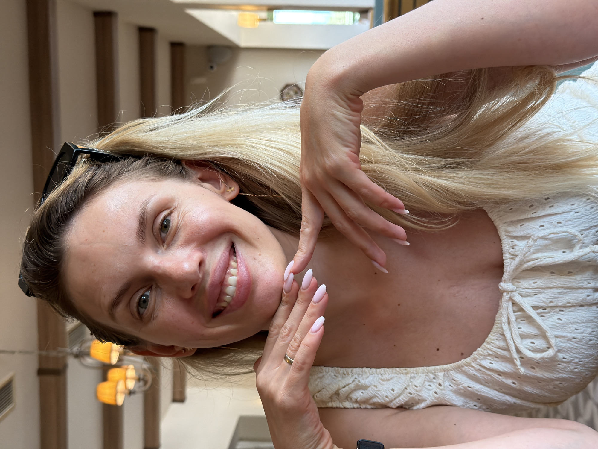 The image shows a medium close-up, selfie-style photograph of a young, blonde woman smiling at the camera. She is wearing a white, eyelet-patterned top with a drawstring at the neckline. Her hands are clasped beneath her chin, and she has long, light-pink, almond-shaped nails. A thin gold band is visible on her ring finger. She has a small gold star earring in her left ear. Her hair is long and blonde, pulled back from her face with a black clip.

The background appears to be an indoor setting, possibly a restaurant, with wooden beams on the ceiling and orange-shaded lamps hanging. The lighting is warm and natural. The overall impression is casual and relaxed. The location is identified as Bigor, Montenegro.