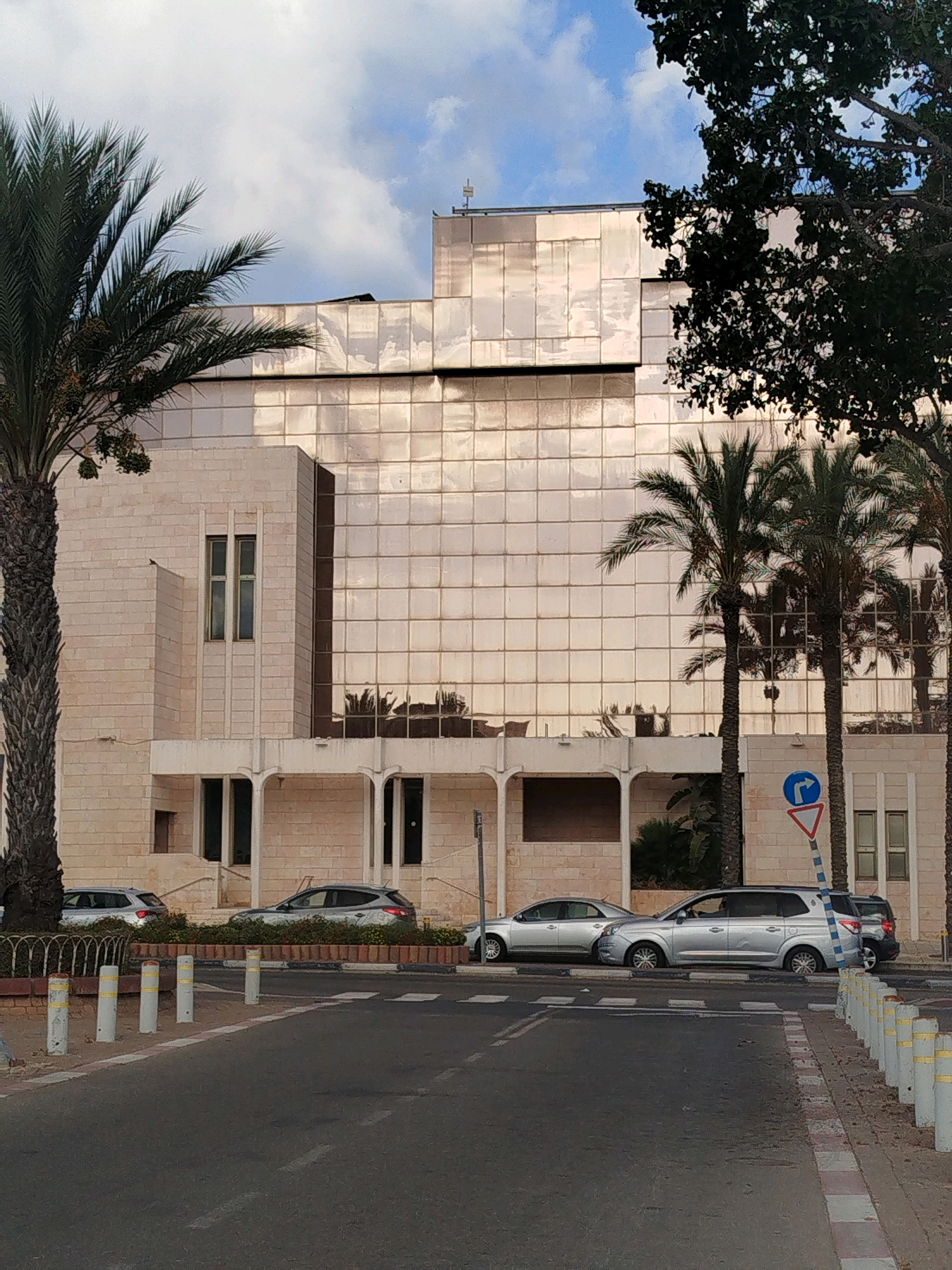 The image shows a street scene in Ashkelon, Israel. In the foreground, a gray asphalt road is visible, with a white and red striped curb on the right side. Several white bollards with yellow reflective stripes are spaced along the curb. A pedestrian crossing with white stripes is marked on the road.
In the middle ground, a row of silver cars is parked along the side of the road. Behind the cars, there is a low, decorative planter box with reddish-brown soil and sparse green foliage.
The background features a large, modern building with a facade of light-colored stone on the lower levels and a reflective, glass-paneled upper section. The glass reflects the sky and surrounding palm trees. There are several tall palm trees on the left and right sides of the image, and a large deciduous tree with dark green foliage is visible on the upper right.
Two windows with dark frames are visible on the stone portion of the building on the left. A set of arched architectural elements is present above the planter box, supporting the upper part of the building. A blue and white directional sign with a blue arrow pointing right and a red triangle with a black border is mounted on a pole near the right side of the building. The sky above is partly cloudy.