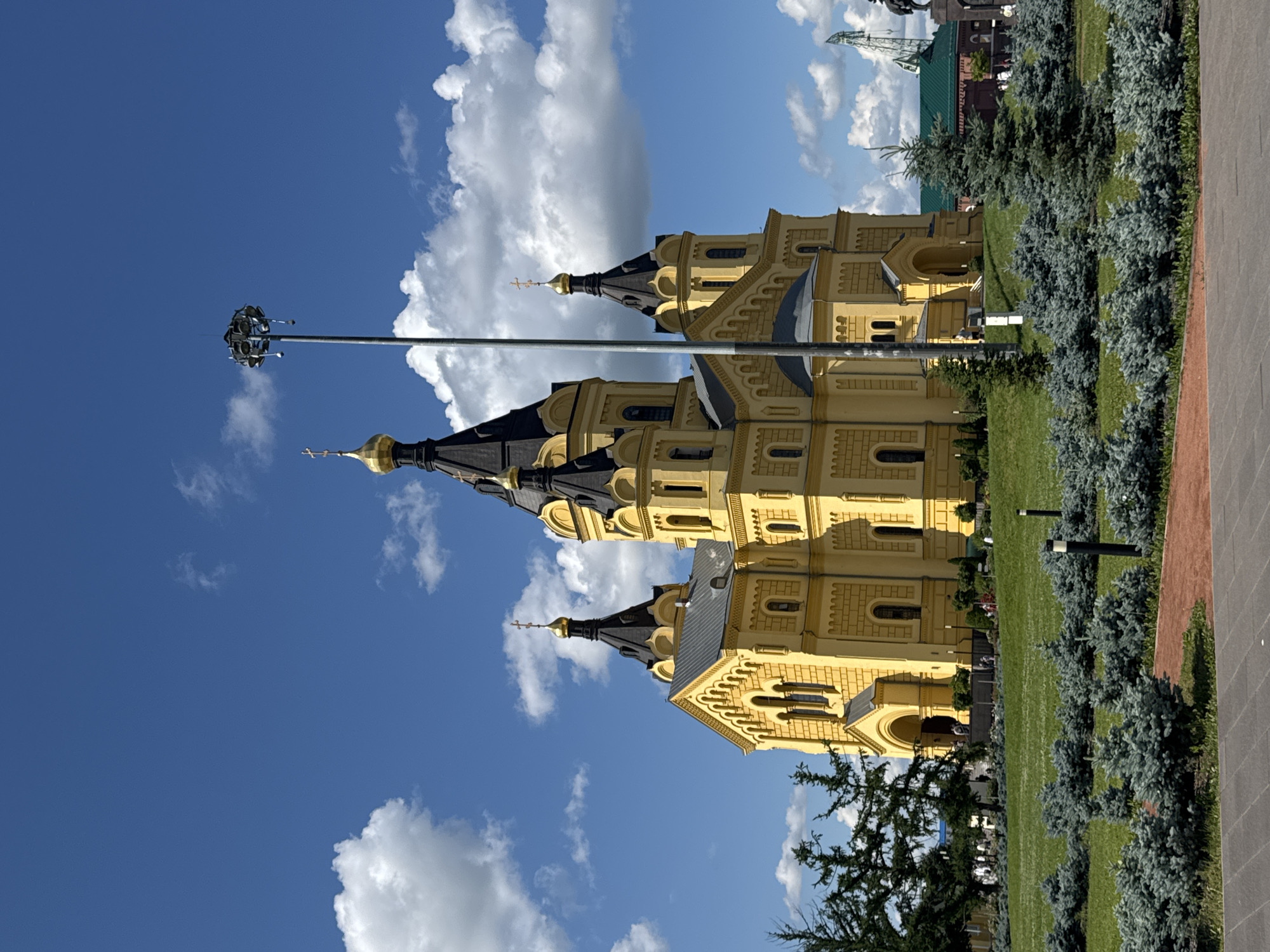 The image depicts the Alexander Nevsky Cathedral in Nizhny Novgorod, Russia, on a sunny day with scattered clouds. The prominent yellow cathedral features multiple black domes topped with golden crosses. In the foreground, manicured green grass is interspersed with low-lying blue-green evergreen shrubs and several modern lamp posts. A tall metal pole with lighting fixtures stands to the right of the cathedral, partially obscuring it. In the background, a section of a green roof and a tall construction crane are visible, along with distant trees and other structures. A few people are seen on the steps and grounds of the cathedral, suggesting a public space. The overall scene is a clear, well-lit view of a significant religious landmark and its surrounding urban environment.
