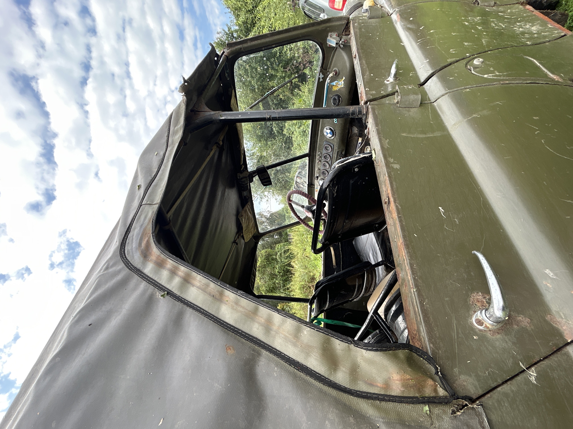 The image captures an exterior view of an old, olive-drab military-style vehicle, possibly a UAZ, parked outdoors in a grassy area. The vehicle appears weathered and worn, with visible rust spots and scratches on its body. The canvas roof is torn and frayed in several places, revealing the metal frame beneath and showing signs of age. The driver's side door handle is also rusty.
Through the open side window and the damaged windshield, the interior of the vehicle is partially visible. It contains two black seats, a steering wheel with a red rim, and a dashboard with various gauges and knobs. The dashboard also has a few stickers, one appearing to be a BMW logo and another with a cartoon character.
The vehicle is situated amidst lush green foliage and trees in the background. The sky above is partly cloudy with white cumulus clouds against a blue sky. To the right of the vehicle, a portion of another vehicle, possibly a silver or light-colored car, is visible, suggesting it is parked nearby. The overall setting appears to be a rural or semi-rural environment. The image does not contain any information that definitively places it in the city of Калицино, Russia.