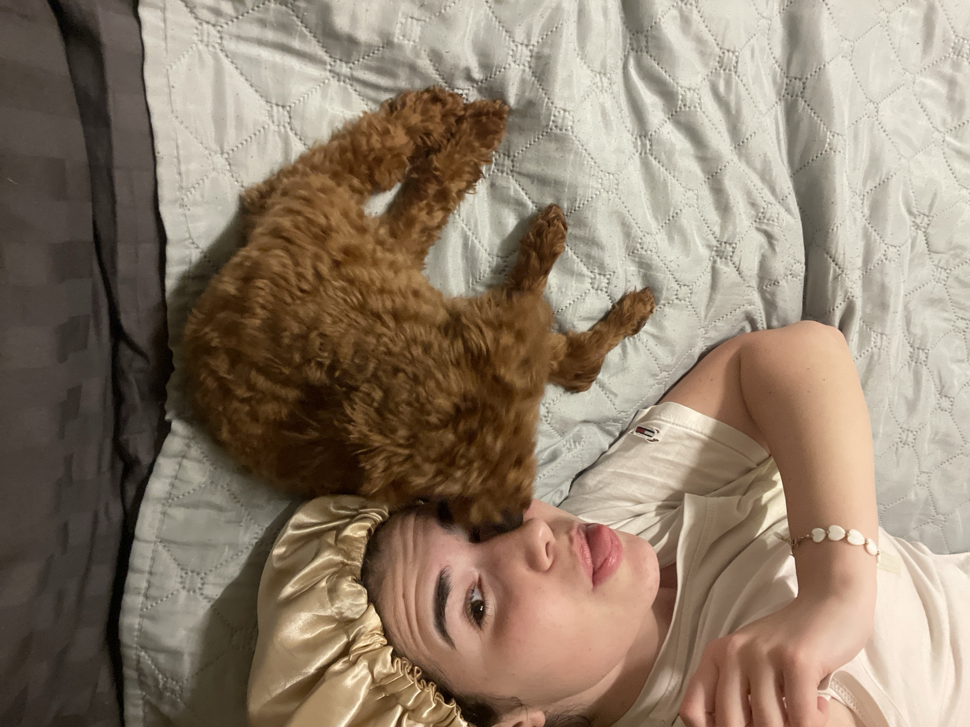 A high-angle shot depicts a young woman lying on a bed alongside a brown poodle. The woman wears a white t-shirt and a beige satin bonnet. A bracelet adorns her left wrist. The poodle is positioned near her head, seemingly resting on her face. The bed is covered in grey bedding. No evidence is present to confirm that this image was taken in Sofia, Bulgaria.