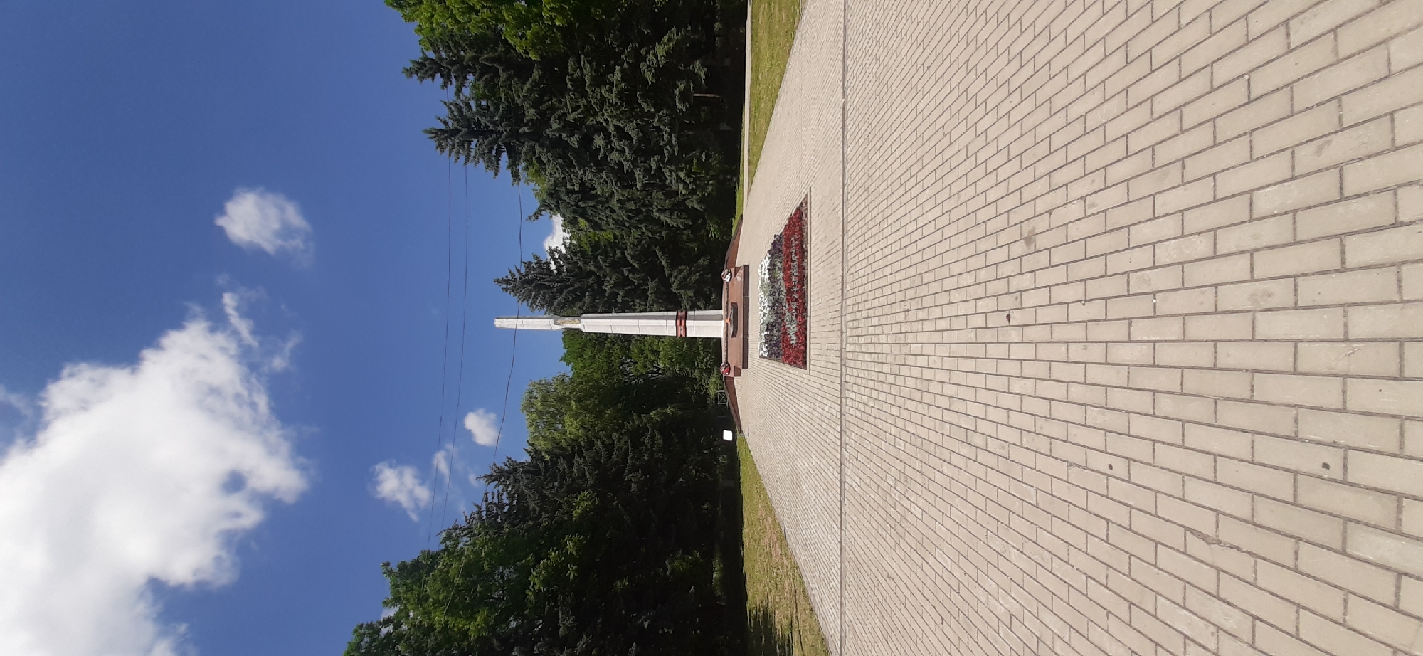 A vertical, eye-level, full shot captures a monument in the city of Yessentukskaya, Russia, on a sunny day. The monument, a tall, white obelisk with red accents, stands at the end of a wide, paved walkway. At the base of the monument, a rectangular flowerbed is filled with red and white flowers, and a perpetual flame burns. The walkway, composed of rectangular paving stones, extends towards the viewer. Lush green trees, including firs, surround the monument and walkway, creating a park-like setting. The sky is a bright blue with scattered white clouds.
