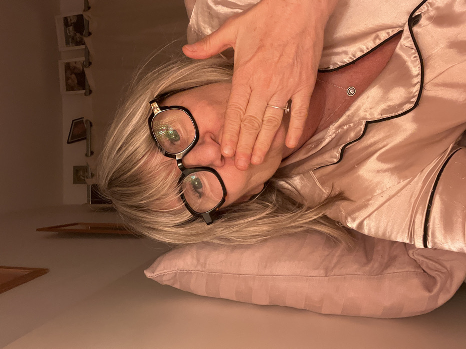A woman with blonde hair, wearing black-rimmed glasses and pale pink satin pajamas with black piping, lies on her side on a matching pillow. Her hand is covering her mouth. A ring is visible on her finger and a necklace is seen on her chest. In the background, there are framed photos hanging on a wall.