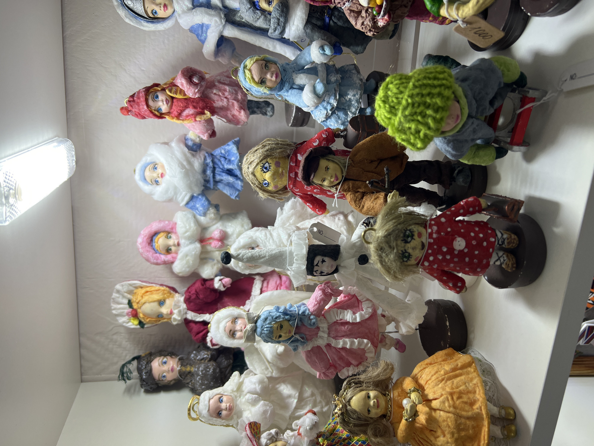 The image shows a shelf displaying a collection of handmade dolls. The dolls are diverse in appearance, attire, and materials. Some are dressed in winter outfits, while others wear more casual clothing. Some appear to be riding sleds, and they are all mounted on stands. The lighting suggests the scene is indoors, and the style of the dolls indicates a craft or souvenir shop in a location such as Chaikovsky, Russia, based on the likely cultural origin of the items.