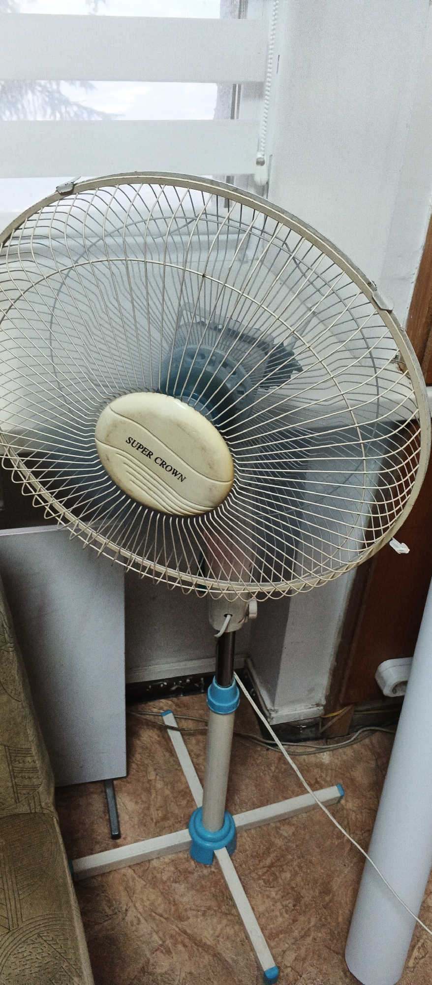 The image shows a close-up of a white electric fan with a blue and white base. The fan has a circular metal grill with a cream-colored oval center that reads "SUPER CROWN". The fan is on a stand with a telescopic pole, adjustable to different heights. The fan is positioned indoors, with a window with horizontal blinds visible in the background. The flooring appears to be tiled. The scene is an interior, likely a residential setting, in the city of Smila, Ukraine.