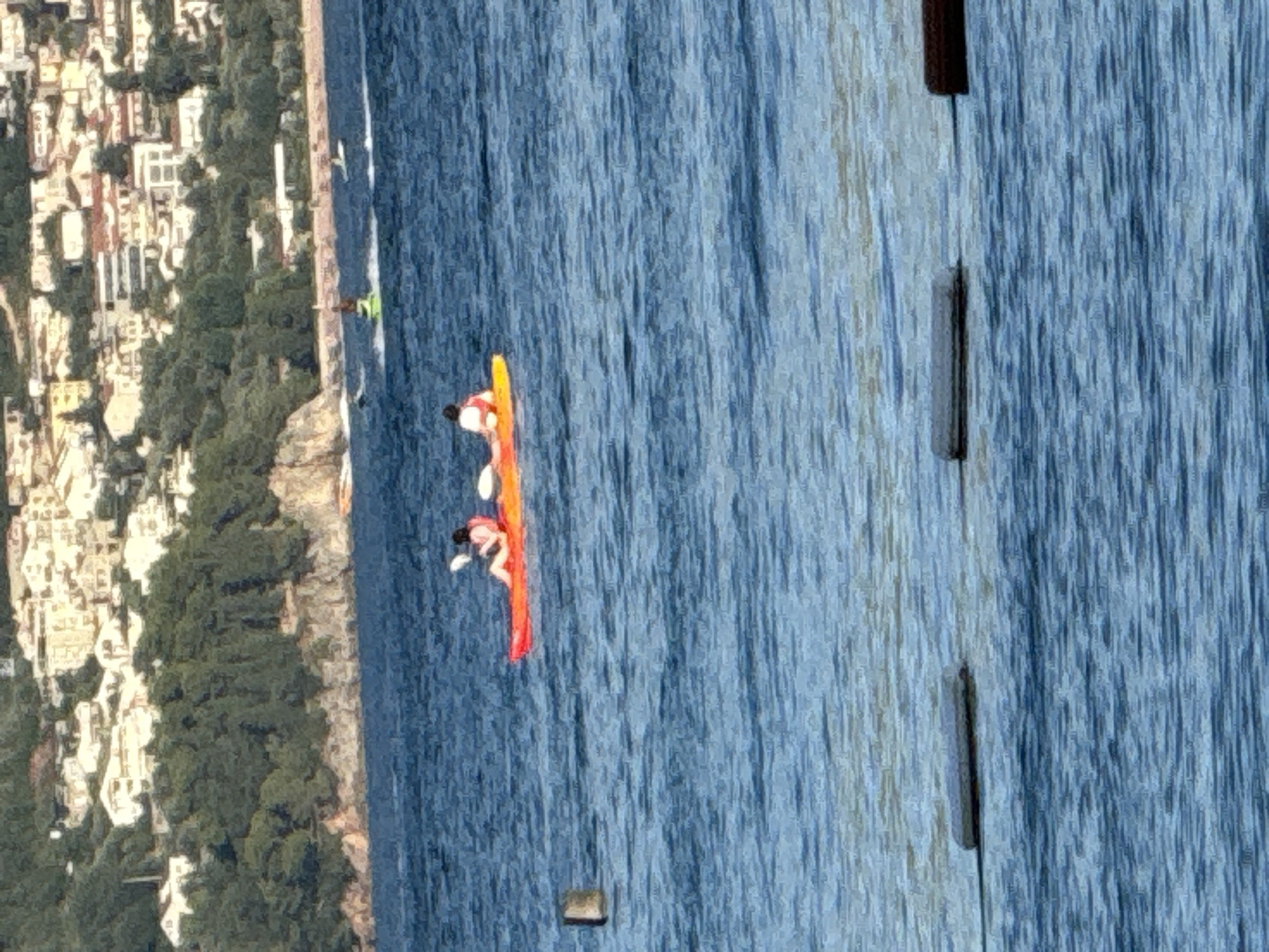 Two people are in a bright orange kayak on the blue water. The person in the front is wearing a white vest and is holding a paddle. The person in the back is wearing a red vest. They are positioned in the middle of the frame. In the background, there is a hill with buildings and trees, likely a coastal city. In the foreground, there are buoys on the water, suggesting a marked area. The water is choppy, indicating some wind or movement.