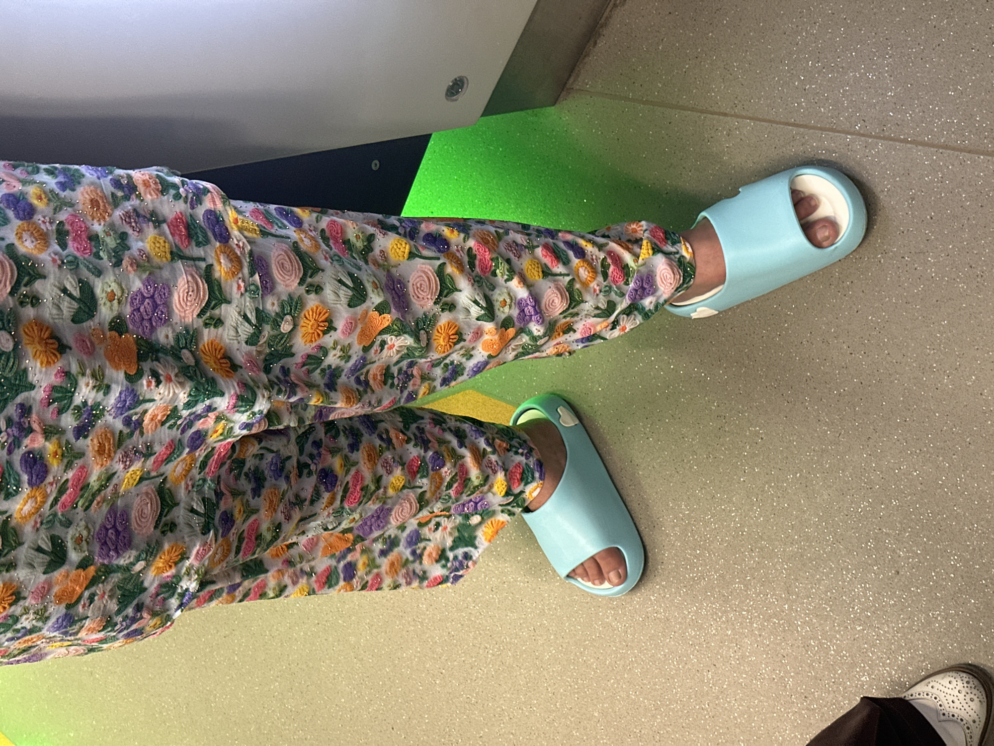 The image shows a person's legs and feet. They are wearing a floral-patterned garment, possibly pants or a dress, with a white background and colorful embroidered flowers in shades of pink, orange, purple, and yellow. The garment also appears to have glittery accents. The person is also wearing light blue slide sandals. The setting appears to be indoors, with a speckled grey floor. A bright green illuminated area is visible in the background, suggesting some kind of lighting feature or display. Another person's feet are visible in the lower right corner, wearing dark pants and white shoes. The overall impression is casual and possibly taken in a public space with modern design elements. There is no visible text in the image. The lighting and the illuminated green area suggest it could be evening or a place with controlled lighting.