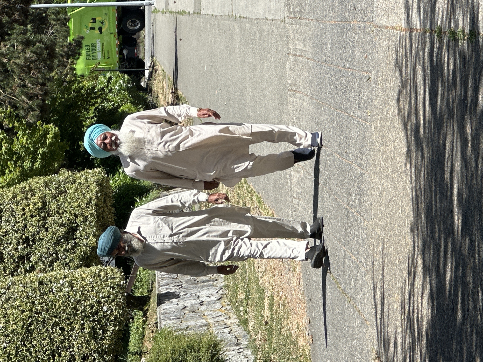 Two men, appearing to be of South Asian descent, walk side-by-side on a paved pathway. The man on the left wears a teal turban and a light-colored tunic and trousers. He has a gray beard and mustache. The man on the right wears a blue turban and a similar light-colored tunic and trousers. He has a white beard and mustache. Both men are wearing dark-colored shoes. Behind them, on the right side of the image, is a bright green garbage truck with text that reads "FUELED BY YOUR FOOD SCRAPS." The background includes lush green trees and a stone wall on the left. The overall setting appears to be an outdoor park or pathway in a city. The garbage truck is a distinct identifier that it belongs to the City of Surrey, Canada, as this is part of their organic waste collection program.
