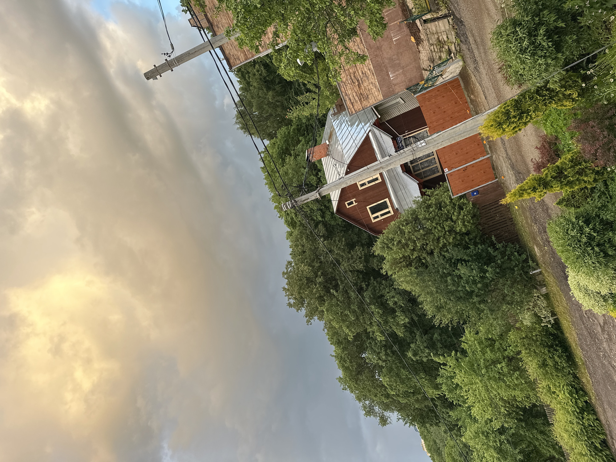 The image depicts a residential area in Nizhny Novgorod, Russia. A red wooden house with a silver roof is the central subject, nestled among trees and adjacent to a road. Power lines run across the frame, connected to a utility pole near the house. The sky is overcast with a mix of light and dark clouds. Another building with a brown roof is partially visible in the background. The environment includes trees, foliage, and a narrow road with a gravel surface.