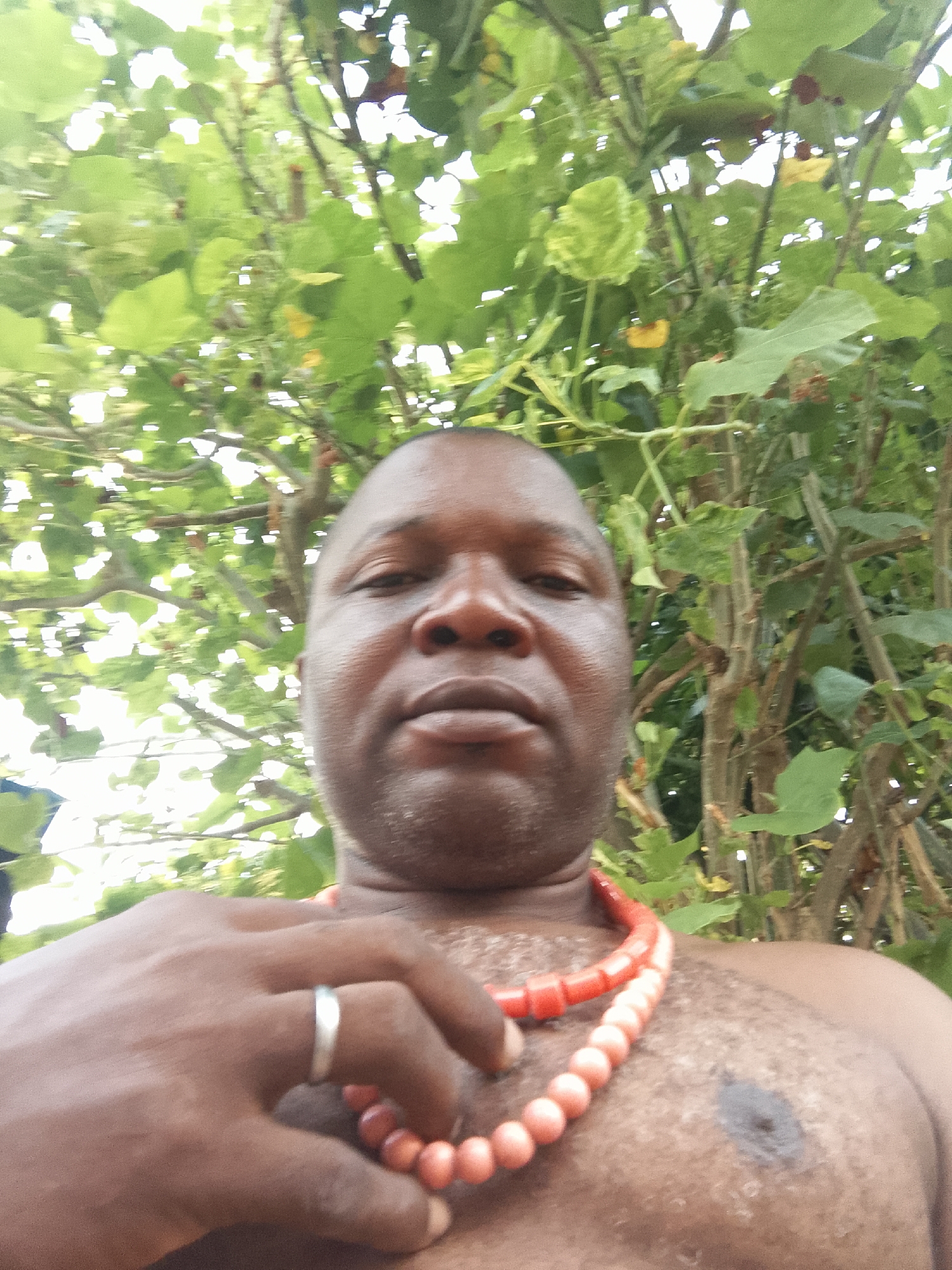 A medium shot depicts a dark-skinned man with a shaved head. He is shirtless, wearing a red beaded necklace, and a silver ring on his left ring finger. He appears to be standing outdoors, with foliage visible behind him. The location is Unknown, Unknown.