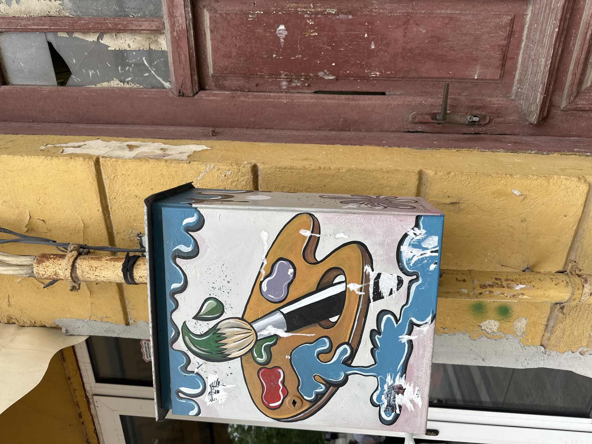 A close-up shot focuses on an artistically painted mailbox affixed to a weathered yellow wall. Above the wall is a maroon window frame, with the paint peeling and some glass panes missing. The mailbox is decorated with a colorful paint palette and a paintbrush, outlined in black and accentuated with blue, white, and gray swirling patterns. Rust is visible on a pipe running horizontally across the wall near the mailbox. In the background, a modern white window is partially visible. The scene suggests a location in a possibly historic area of Vidin, Bulgaria, given the combination of older, weathered building elements and the more modern window in the background.