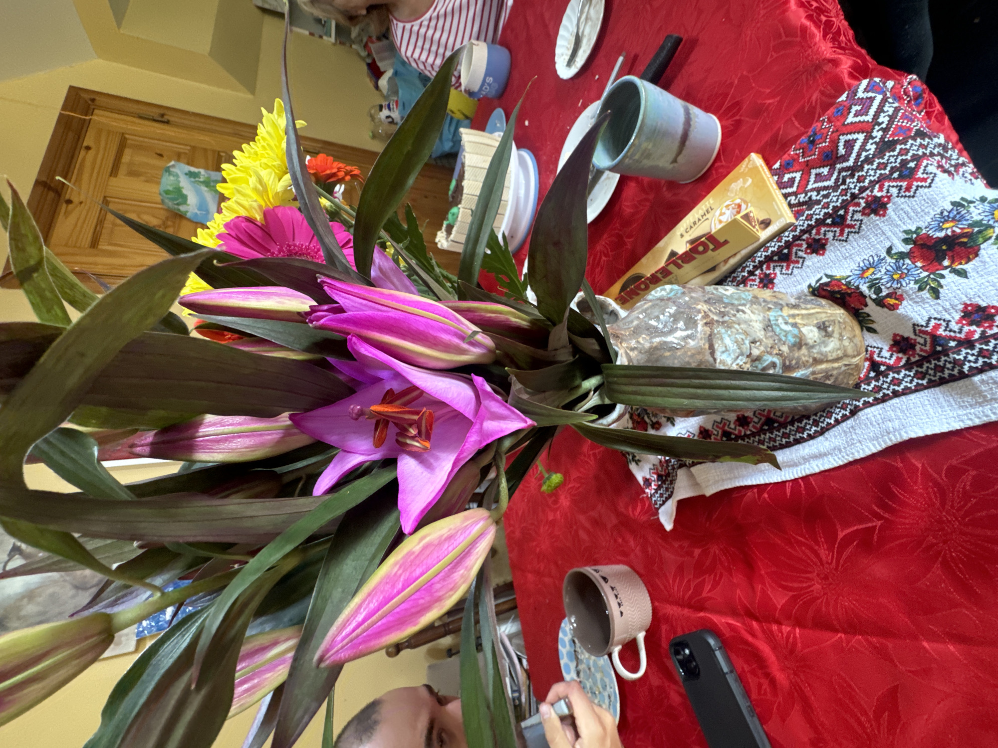 The image depicts a table set for a meal, likely a celebration, with a prominent floral arrangement as the centerpiece. The table is covered with a bright red tablecloth, which has a subtle embossed floral pattern. To the left, a person's head and shoulder are visible, suggesting they are seated at the table, possibly looking at something off-camera or engaged in a conversation. A smartphone lies on the table to the left of the floral arrangement.
The floral arrangement itself is lush, featuring large pink lilies and vibrant yellow and pink gerbera daisies, accompanied by dark green foliage. The lilies are in various stages of bloom, with some fully open and others still in bud. They are contained within an ornate, textured vase.
On the table, to the right of the centerpiece, is a partially visible cake on a white cake stand, decorated with green frosting and what appear to be wafer cookies around the sides. Several plates, some with remnants of food, are also present, along with cutlery and a coffee mug. A blue and white ceramic mug with the inscription "MOR." is visible further to the right. A box of Toblerone chocolate is placed near the vase. A decorative textile with embroidered floral and geometric patterns lies beneath the vase and partially extends across the table.
In the background, a wooden door with a brass handle is visible, and a portion of a wall painted yellow. The overall ambiance suggests an indoor gathering, possibly a home setting. The presence of a cake and celebratory flowers indicates a special occasion. While the location is stated to be in Waterford, Ireland, there are no specific visual cues within the image to pinpoint a precise neighborhood or landmark within the city. The scene is well-lit, likely by natural light or a combination of artificial and natural light sources.