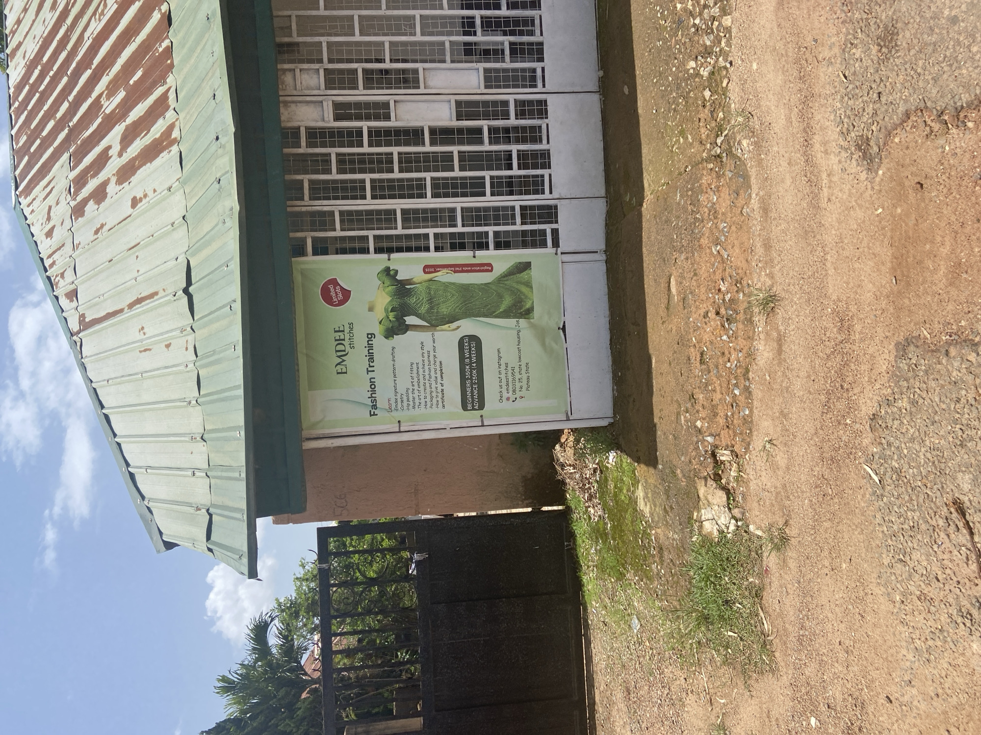 The image shows the exterior of a shop with a corrugated metal roof, some of which is rusted. A green banner advertises "EMDEE stitches Fashion Training" and lists the curriculum, including "Learn - Emdee signature pattern drafting," "Hip padding," and "Master the art of fitting." It also mentions "The art of embellishment," "How to create and achieve any style," and "Packaging and Fashion business," concluding with "How to give value to your worth." The banner also states "BEGINNERS 350K (8 WEEKS)" and "ADVANCE 250K (4 WEEKS)," with a "Limited Slots" designation. Contact information is provided, including an Instagram handle "@emdeestitches," a phone number "08036169922," and a location "No. 25, state lowcost housing, Jos, Plateau State." A tall green dress is depicted on the banner. To the left of the banner is a large black metal gate, and a light-colored building with a patterned window covering is visible to the right. The ground is a mix of dirt and gravel with some patches of grass. The sky above is blue with scattered clouds.