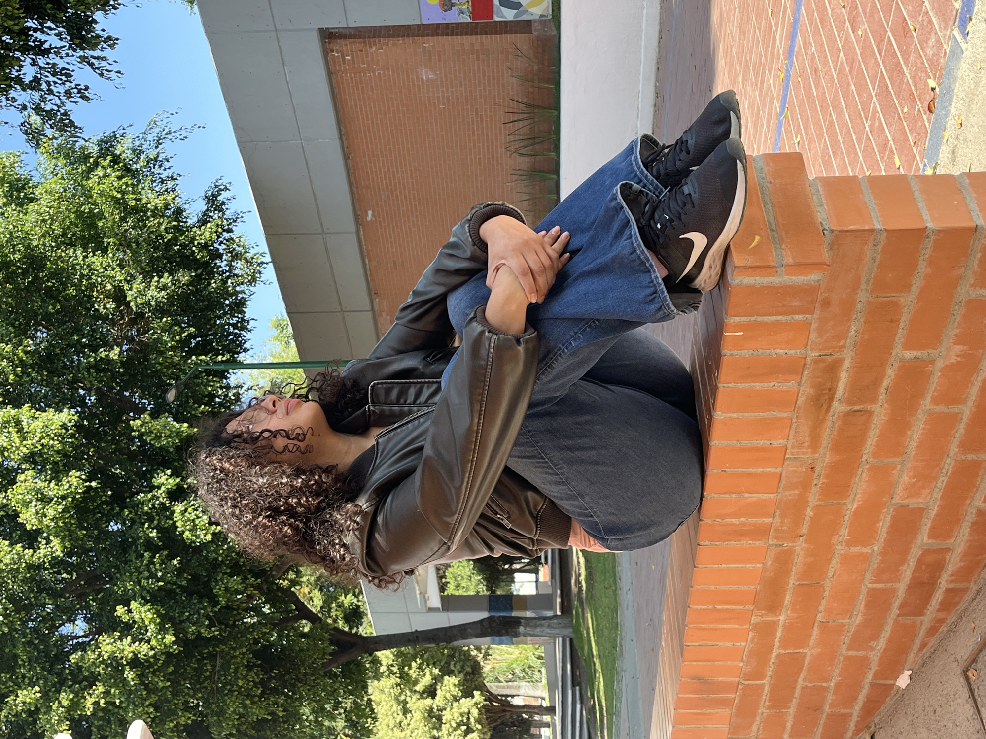A young female, likely in her late teens or early twenties, with dark, curly hair and eyeglasses, is seated in a profile view on a reddish-orange brick wall. She is positioned facing towards the right side of the frame, looking upwards and to her right. She wears a dark brown leather bomber jacket over a light pink undergarment, blue jeans with flared cuffs, and black Nike athletic shoes featuring white swoosh logos and white soles. Her legs are drawn up, with her knees bent, and her hands are clasped together over them.
The setting is an outdoor area consistent with a university campus, specifically identifiable by architectural elements such as the distinct concrete walls and painted pillars, as the Benemérita Universidad Autónoma de Puebla (BUAP) Ciudad Universitaria campus in Puebla, Mexico. The background features lush green trees under a clear blue sky. Behind the subject, a light gray concrete wall with horizontal panel lines is visible. To the right, a segment of a colorful mural or abstract artwork is present on a lower wall section, in front of which small green plants grow. The ground visible in the foreground is paved, exhibiting a blue painted line to the right of the subject and scattered fallen leaves. The scene is brightly illuminated by natural sunlight, indicating clear weather.