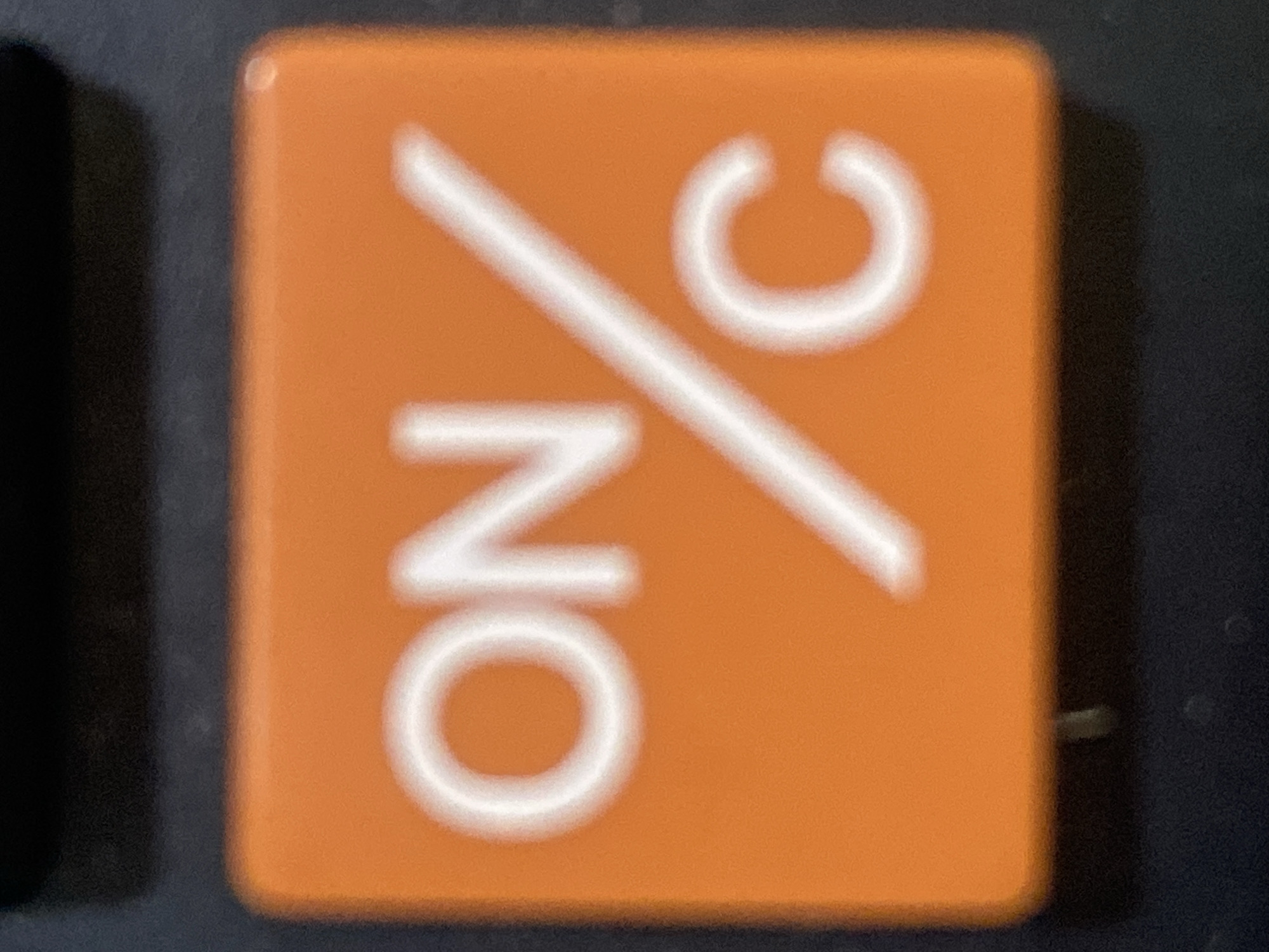 The image shows a close-up, slightly blurry, eye-level view of an orange calculator button. The button has white text and symbols on it. The text reads "ON" on the left side, and "C" on the right side, with a diagonal slash between them. The button is on a dark, textured surface, likely the rest of the calculator. There are no people or other environmental details visible. The image does not contain any information related to Rozhny, Ukraine.