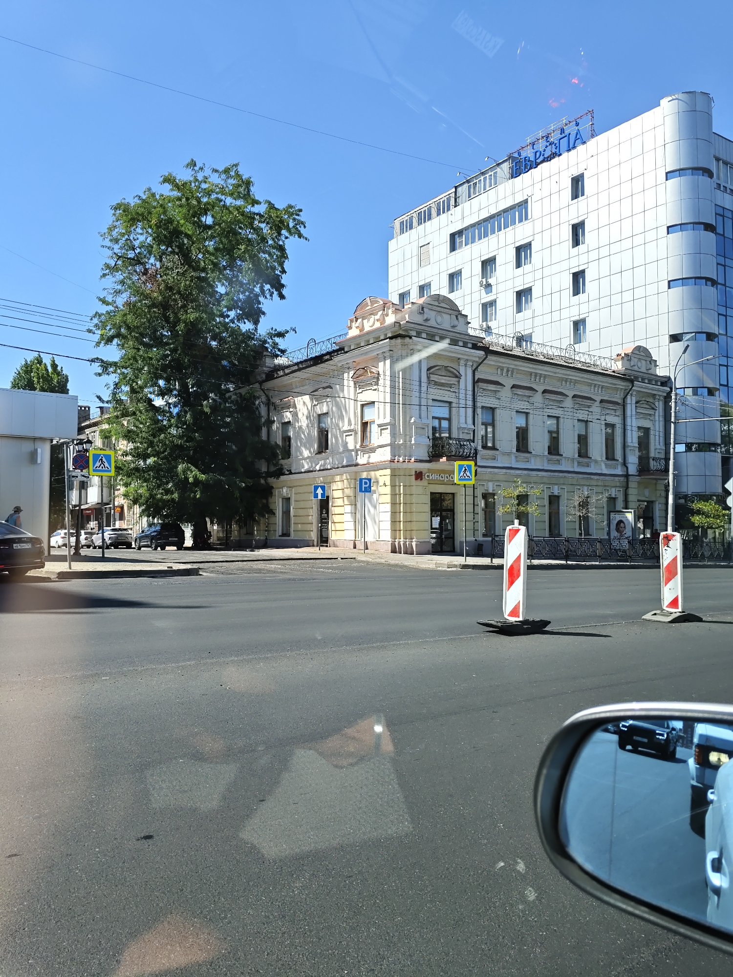 The image depicts a street scene in Rostov-on-Don, Russia, on a clear, sunny day. In the foreground, the asphalt road is visible, with a car's side mirror reflecting other vehicles. Two temporary traffic barriers with red and white stripes are positioned on the road. In the middle ground, a two-story, light-yellow building with ornate architectural details stands to the right of a large, mature tree. A modern, white, multi-story building with a rounded glass facade is visible behind it. Traffic signals, street signs, and parked cars are present. A pedestrian crossing is marked on the road. In the background, a clear blue sky is visible. The overall impression is of an urban environment with a mix of older and modern architecture.