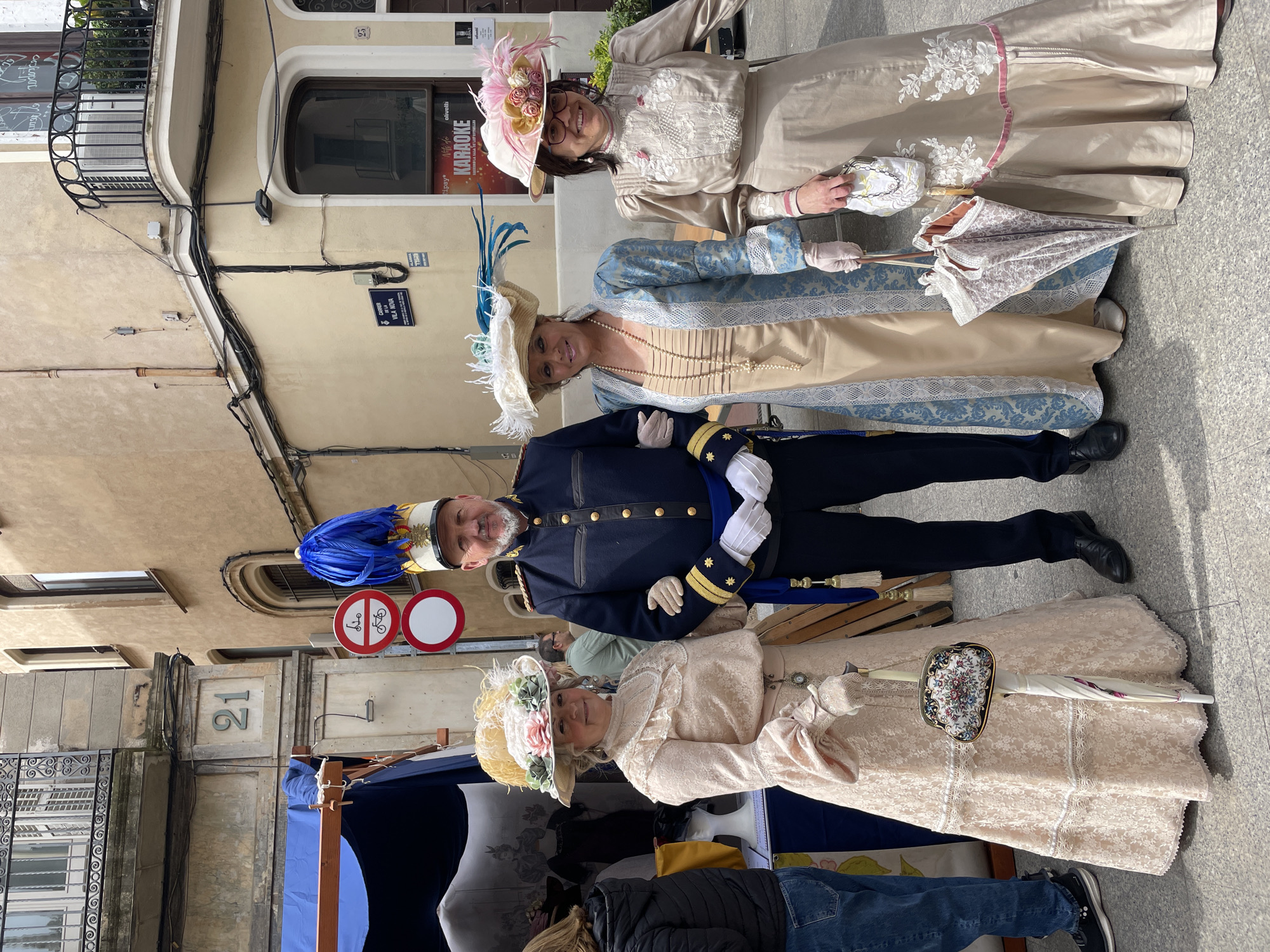 Three women and a man, dressed in period costumes, stand on a paved street in Terrassa, Spain. The women wear hats with feathers and flowers and carry umbrellas and purses. One man is in a navy blue uniform with a blue feathered hat. A blue canopy is partially visible in the foreground. Building number 21 is shown, as well as a sign reading "Carrer Vila Nova". Another sign indicates "Karaoke". A street sign showing the location is visible. A bicycle sign is also present.