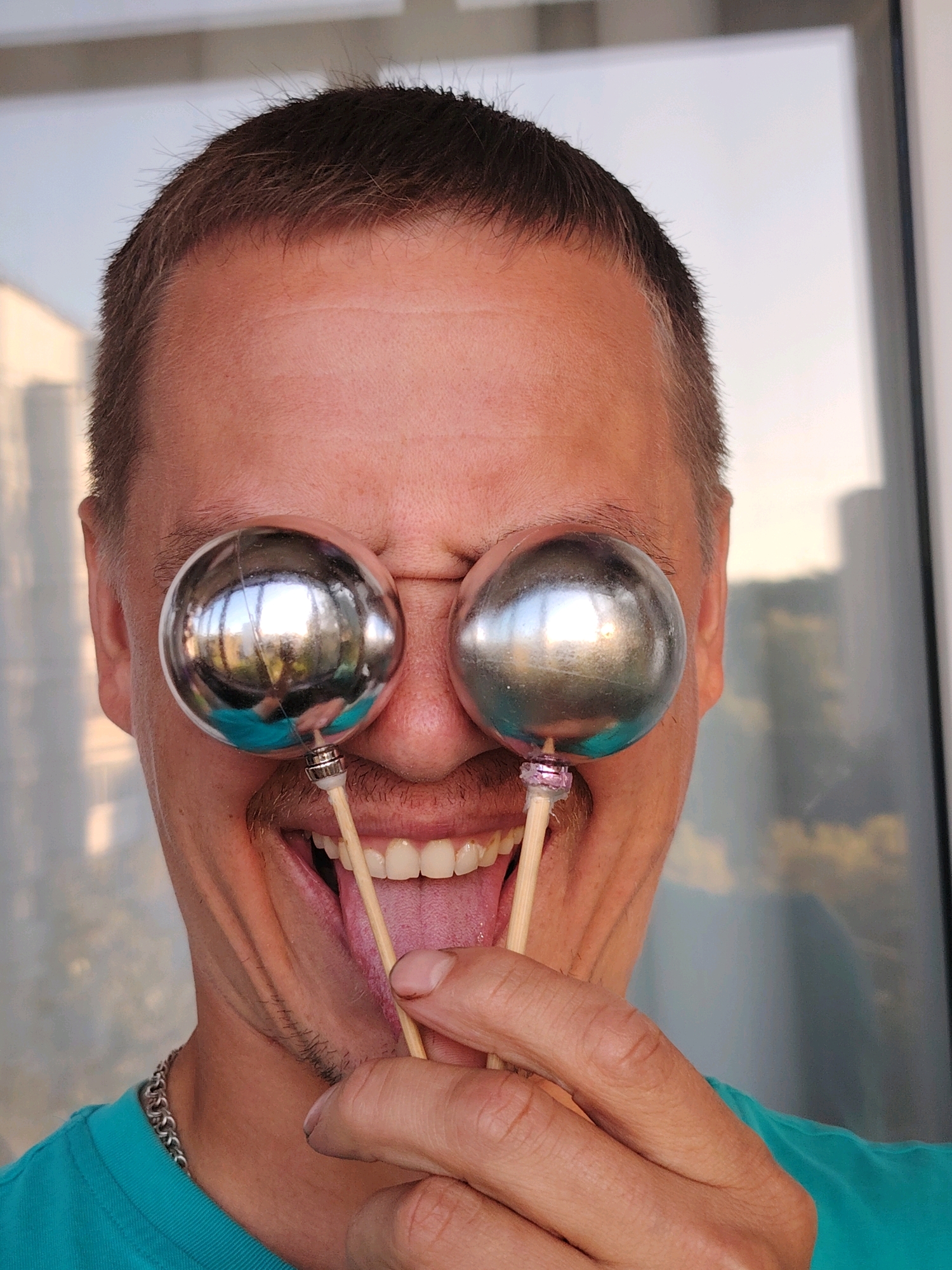 A close-up, eye-level shot shows a man's face from the forehead down, with two shiny, spherical objects held up to his eyes, covering them. The man has short, dark hair with some graying at the temples, fair skin, and a prominent mustache. He is smiling widely, with his mouth open and tongue sticking out, and his eyes are squeezed shut behind the spheres. The spheres appear to be metallic, possibly chrome or aluminum, and are mounted on wooden sticks. One sphere is positioned over his left eye, and the other over his right. A silver chain necklace is visible around his neck, and he is wearing a turquoise t-shirt. The background is blurred, showing a large window or glass door with reflections of buildings and sky, suggesting an indoor environment with an outdoor view. The lighting is bright and natural, casting subtle shadows on the man's face. The scene is set in the city of Chaikovsky, Russia.