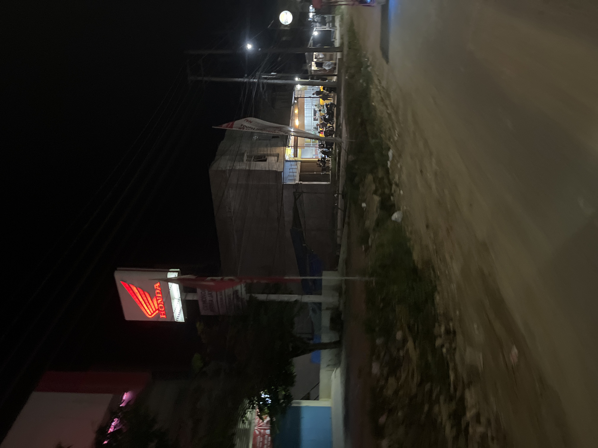 This is a nighttime, outdoor, low-angle shot of a street in Bundar, Indonesia. The scene depicts a Honda dealership. In the foreground, a brightly lit Honda sign with its distinctive wing logo illuminates the dark. To the left, a portion of a building is visible, with some areas appearing to be under renovation or construction. The building's facade is a muted beige, contrasting with the vibrant red of the Honda logo.
Behind the dealership, a street stretches into the distance, lined with more buildings and illuminated by streetlights and the light spilling from the businesses. Several motorcycles are parked in front of the dealership, suggesting it is either closing or has recently closed for the day. Some figures, likely customers or staff, are visible inside the brightly lit establishment, engaged in conversation or activity. A red and white banner hangs from a flagpole in front of the dealership, with visible text that appears to be in Indonesian, though it is partially obscured by the banner's folds and the angle of the shot. One section of text clearly reads "HONDA." The overall atmosphere is one of a typical Indonesian street scene at night, with the buzz of commerce present despite the late hour. The road surface is visible, appearing to be paved and slightly damp, reflecting the ambient light. The presence of utility poles and wires indicates that the area is well-connected.