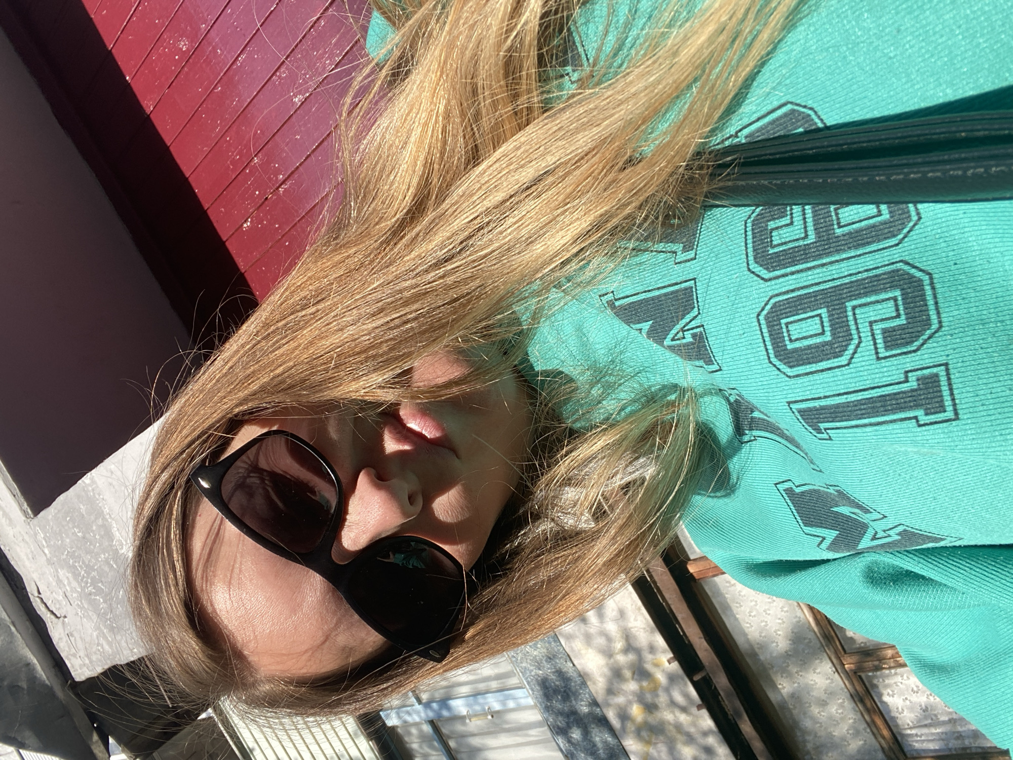 A woman with long, blonde hair and black sunglasses is wearing a turquoise sweatshirt with a dark gray number "96" printed on it. The photo is taken at an angle, with the woman's face slightly tilted to the side. To the left of her face is a white surface with sunlight falling on it, and a red wooden wall is visible in the upper left corner. The background shows a street or outdoor area in the city of Noviy, Russia.