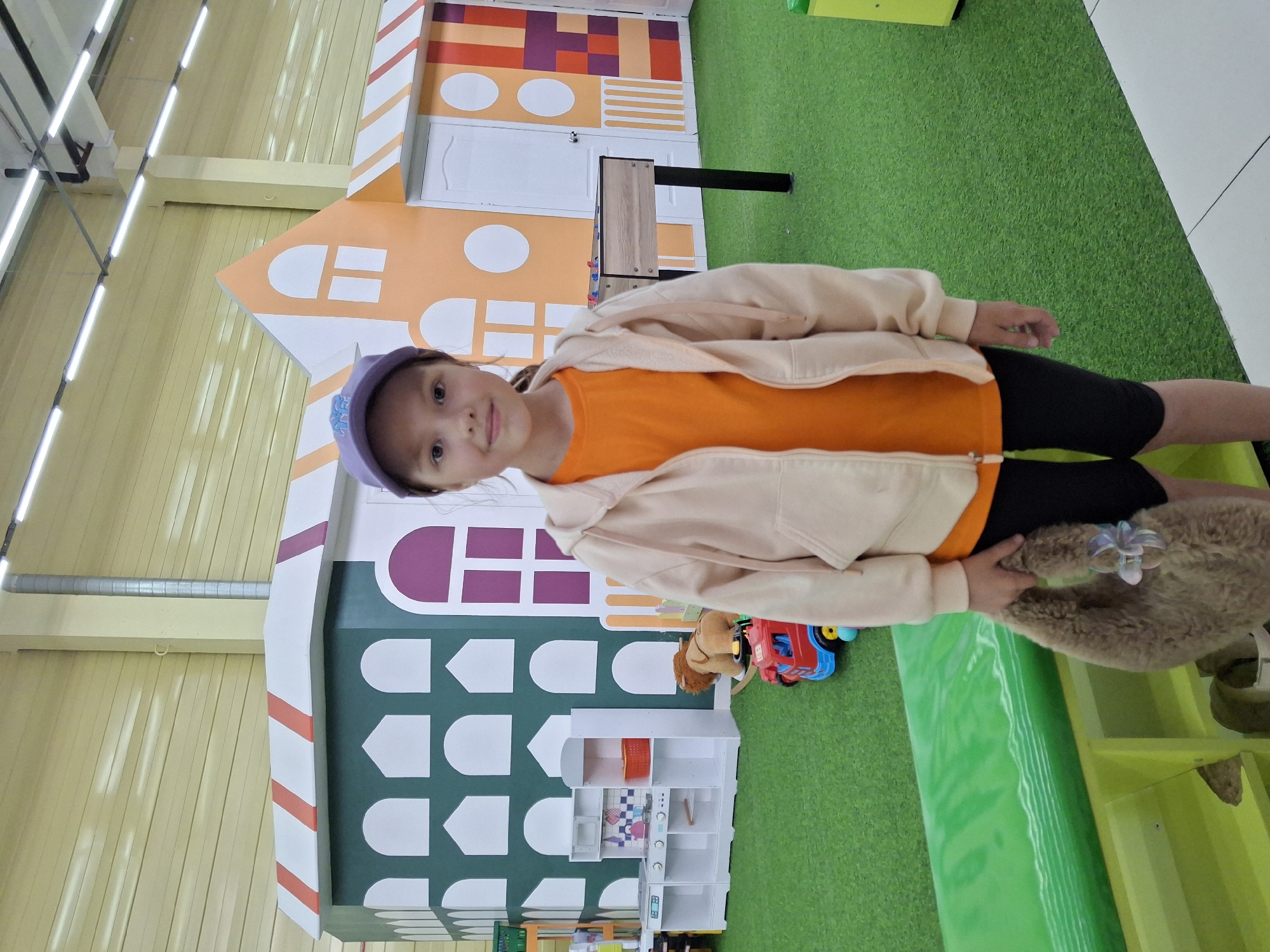 The image depicts a young girl standing in what appears to be an indoor play area. She is wearing a purple baseball cap, a light-colored hooded jacket over an orange t-shirt, and black shorts. She is holding a fluffy, brown bag in her right hand. Behind her, there are colorful, stylized structures resembling buildings, and green artificial turf covers the floor. Scattered on the turf are toys, including a toy kitchen set, a toy truck, and a stuffed lion. In the background, there is a foosball table. The lighting suggests an indoor environment. This scene is consistent with a children's entertainment center, possibly located in the city of Chaikovsky, Russia, given the prompt's context.