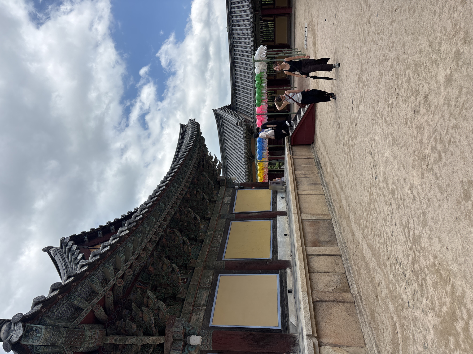 Two women walk across a sandy courtyard in front of a traditional Korean temple building in Gyeongju-si, South Korea. The temple features a dark, tiled roof with ornate eaves and colorful painted decorations. Behind the temple, another building with a similar tiled roof is visible, adorned with strings of colorful lanterns. The sky above is partly cloudy with patches of blue. The woman on the left wears a white tank top and dark pants, carrying a brown shoulder bag. The woman on the right wears a black tank top and dark, flowing pants, holding a black umbrella. Several other people are seen walking up stairs in the background, near the lanterns. The ground is covered in light brown sand, with a low stone wall bordering the temple building.