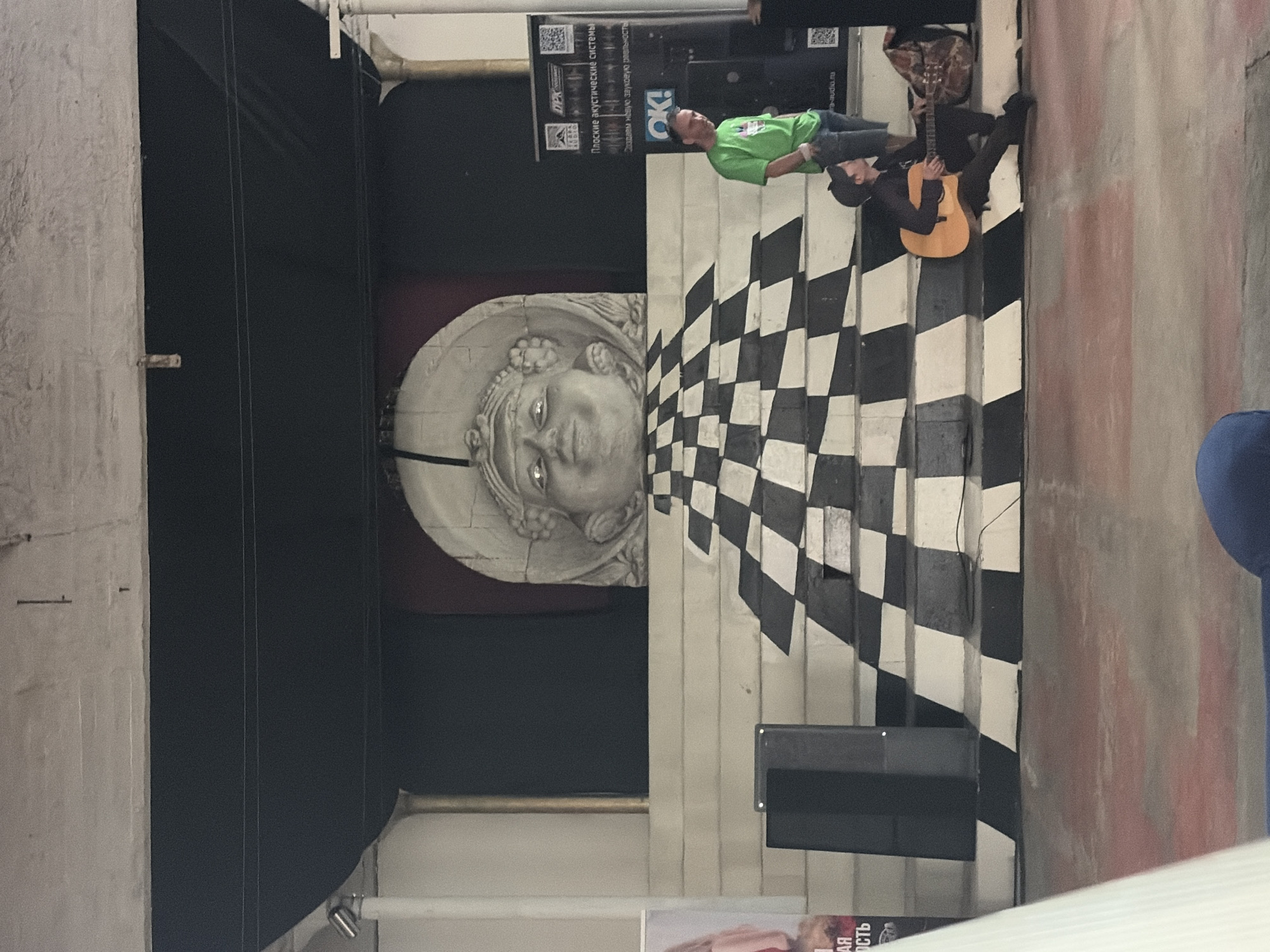 In Il'inskiy Pogost, Russia, a stage area is set up with a large, circular, carved stone face as a backdrop. A checkered black and white pattern ascends the stairs leading to the stage. A man in a green t-shirt and jeans stands on the right side of the stairs, looking towards the left. Below him, a person in dark clothing sits on the stairs playing an acoustic guitar. A guitar case is placed behind the guitarist. Two black speakers are positioned at the base of the stairs on the left side. A large black fabric hangs behind the carved face, and a black banner with text and QR codes is visible on the right. A white banner with pink lettering is partially visible on the far left. The ground in front of the stage is concrete with reddish markings.