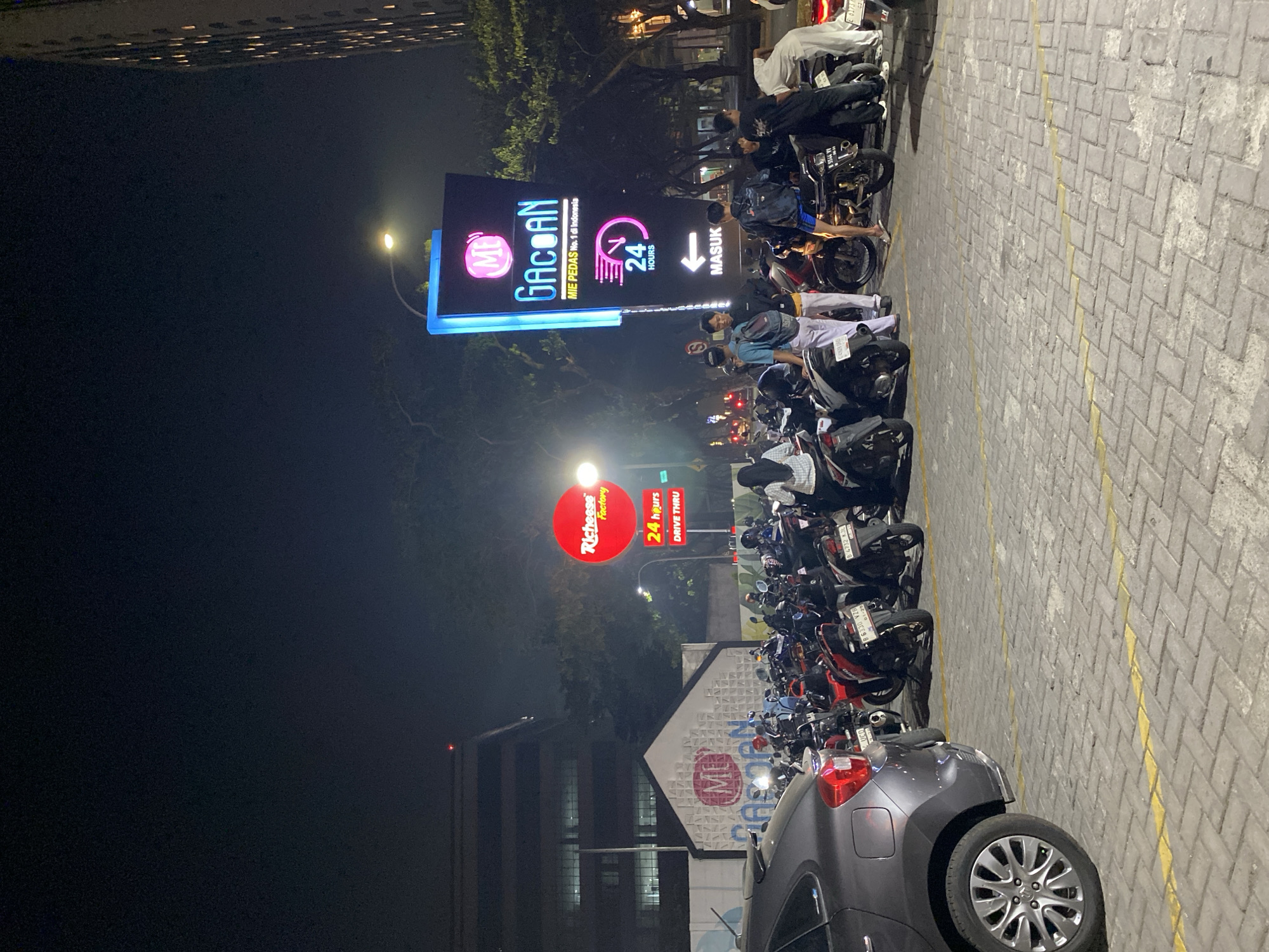 A nighttime scene captures a parking lot in East Jakarta, Indonesia, with a prominent illuminated sign for "MIE PEDAS GACOR" and "Richeese Factory." The "MIE PEDAS GACOR" sign, a tall, vertical structure with blue and purple lighting, advertises "No. 1 di Indonesia" and "24 HOURS." Nearby, a circular red sign for "Richeese Factory" also indicates "24 hours DRIVE THRU." A group of young men are gathered around parked motorcycles, some appearing to be patrons of the establishments. A dark grey car is parked in the foreground on the left, its rear visible. The parking lot is paved with grey interlocking bricks, marked with yellow lines. The background includes a large, dark building with illuminated windows and trees.
