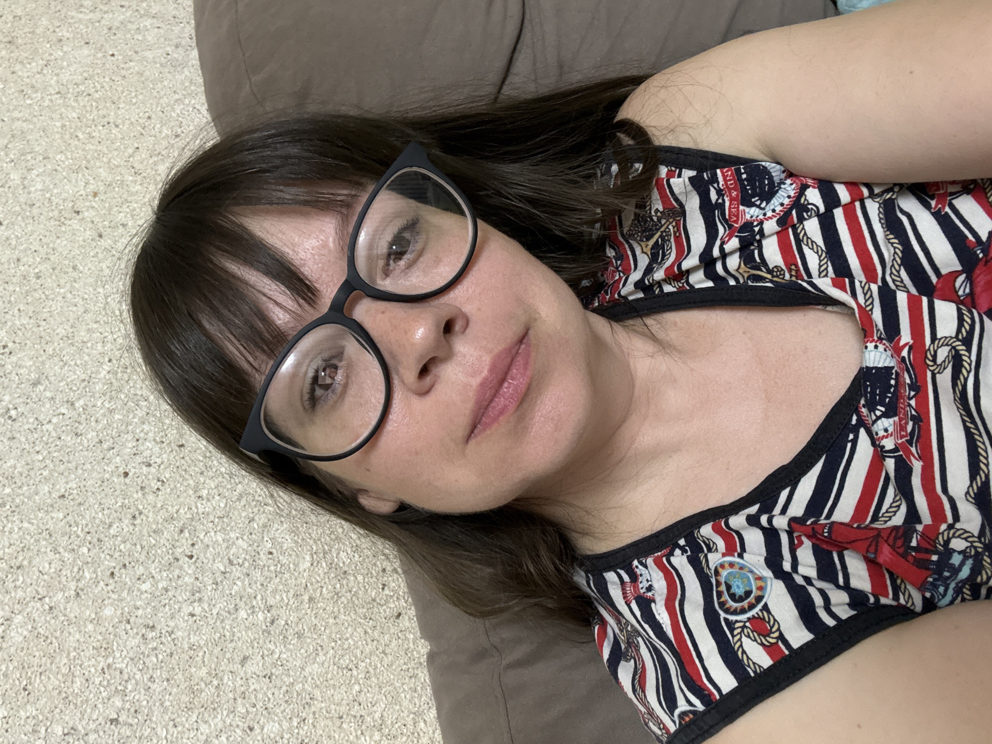 A close-up, overhead shot shows a woman wearing black-rimmed glasses and a sleeveless shirt with a nautical print, featuring red, white, and blue stripes adorned with ropes, anchors, and ships. The woman has dark brown hair with bangs and is looking directly at the camera, her lips slightly parted. She is positioned on a couch, with a textured wall in the background. The specific location within Vetluzhsky, Russia, cannot be determined from the image.