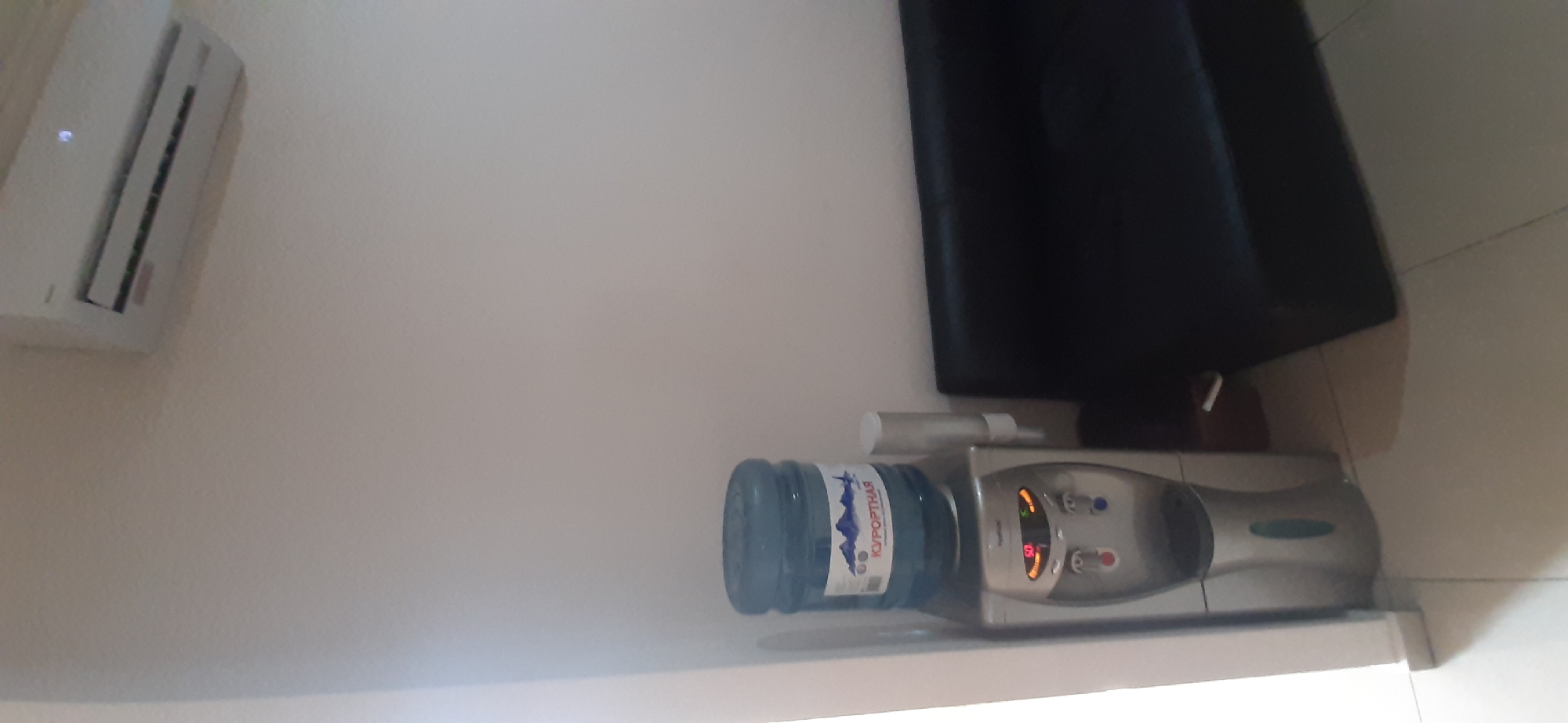 The image shows a water cooler with a large blue water bottle placed on top. The water bottle has a label with text in Cyrillic script, which appears to be "КУРОРТНАЯ". The water cooler is silver and has red and blue buttons, as well as a digital display showing "50". To the left of the water cooler, there is a white air conditioning unit mounted on a light-colored wall. In the background, a dark, possibly black, sofa is visible. The scene appears to be indoors, possibly in a room or lounge area. The lighting suggests it could be daytime, but there are no strong indications of natural light. The overall impression is of a functional space, likely a hotel room, office, or common area in a resort, given the "КУРОРТНАЯ" label which translates to "resort" or "spa" in Russian, and the location context of Yessentuki, Russia, a well-known spa town. There are no people visible in the image.