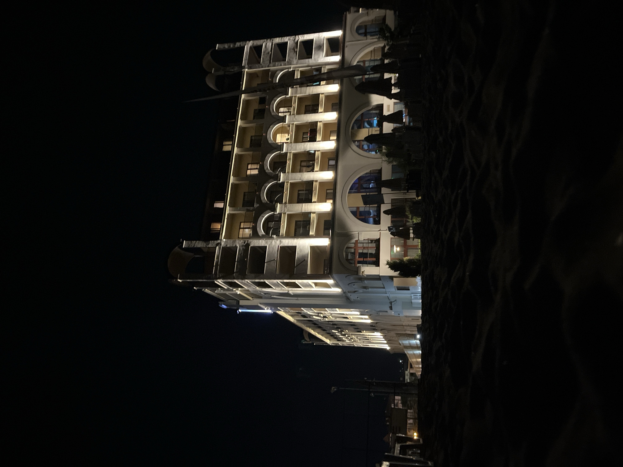 A low-angle shot captures a large, illuminated building at night, situated on what appears to be a sandy area in the city of Obzor, Bulgaria. The building is multi-storied and features a light-colored facade, with numerous windows and balconies. It is brightly lit by exterior lights that accentuate its architectural details, including arched doorways on the lower levels and decorative elements on the roofline. In front of the building, several dark, closed umbrellas are visible, suggesting an outdoor seating area. The foreground is dominated by dark, textured sand, creating a sense of depth and foreground interest. The sky is a deep black, indicating nighttime.