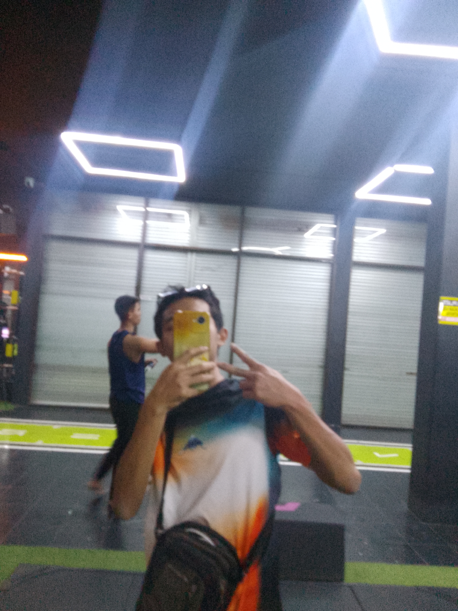 The image captures a selfie taken by a young man in a gym. He is in the foreground, holding a yellow smartphone and making a peace sign with his fingers. He is wearing a t-shirt with a colorful abstract design. Behind him, a second young man in a blue tank top is visible, also reflected in a mirror. The gym environment features modern lighting with rectangular illuminated fixtures on the ceiling. The walls appear to be metallic roll-up doors, and the floor has green markings. There is no specific information in the image to indicate the location is Trenggalek, Indonesia.