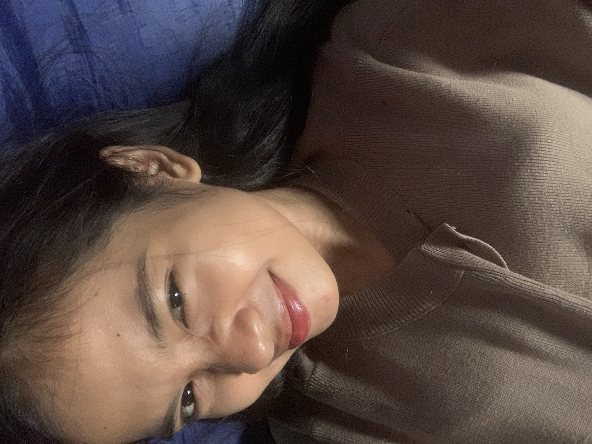 A close-up shot features a young woman with dark hair and light brown skin, lying down on a blue surface, likely a bed or sofa. She is looking towards the camera with a playful expression, one eye winking. Her lips are painted with a glossy, reddish-pink lipstick. She is wearing a brown, ribbed knit sweater. The lighting appears to be artificial and somewhat dim, highlighting the sheen on her skin. The overall impression is that of a casual, candid selfie taken indoors. There is no information available to determine the specific location in Sungai Meriam, Indonesia.