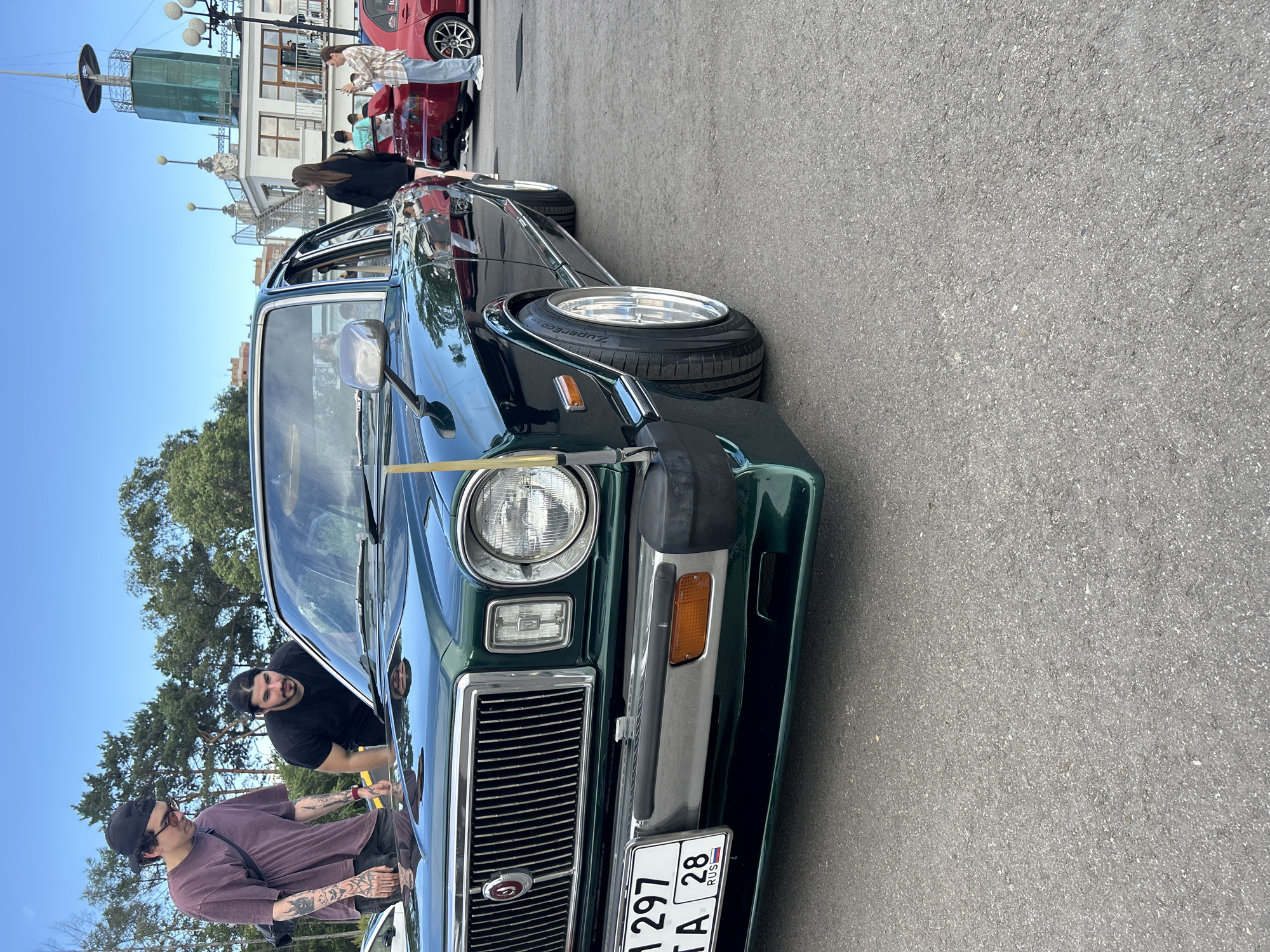 In Khabarovsk, Russia, a dark green vintage sedan is parked on asphalt. Two men, one wearing a purple t-shirt and black baseball cap, and the other in a black t-shirt, are examining the car. In the background, several other cars, including a red hatchback, are visible. People are milling about in the background, with a distinctive building featuring a lighthouse-like structure and a green dome visible in the distance. The sky is clear and blue, suggesting a sunny day. The license plate on the front of the sedan reads "M 1297 TA 28 RUS".