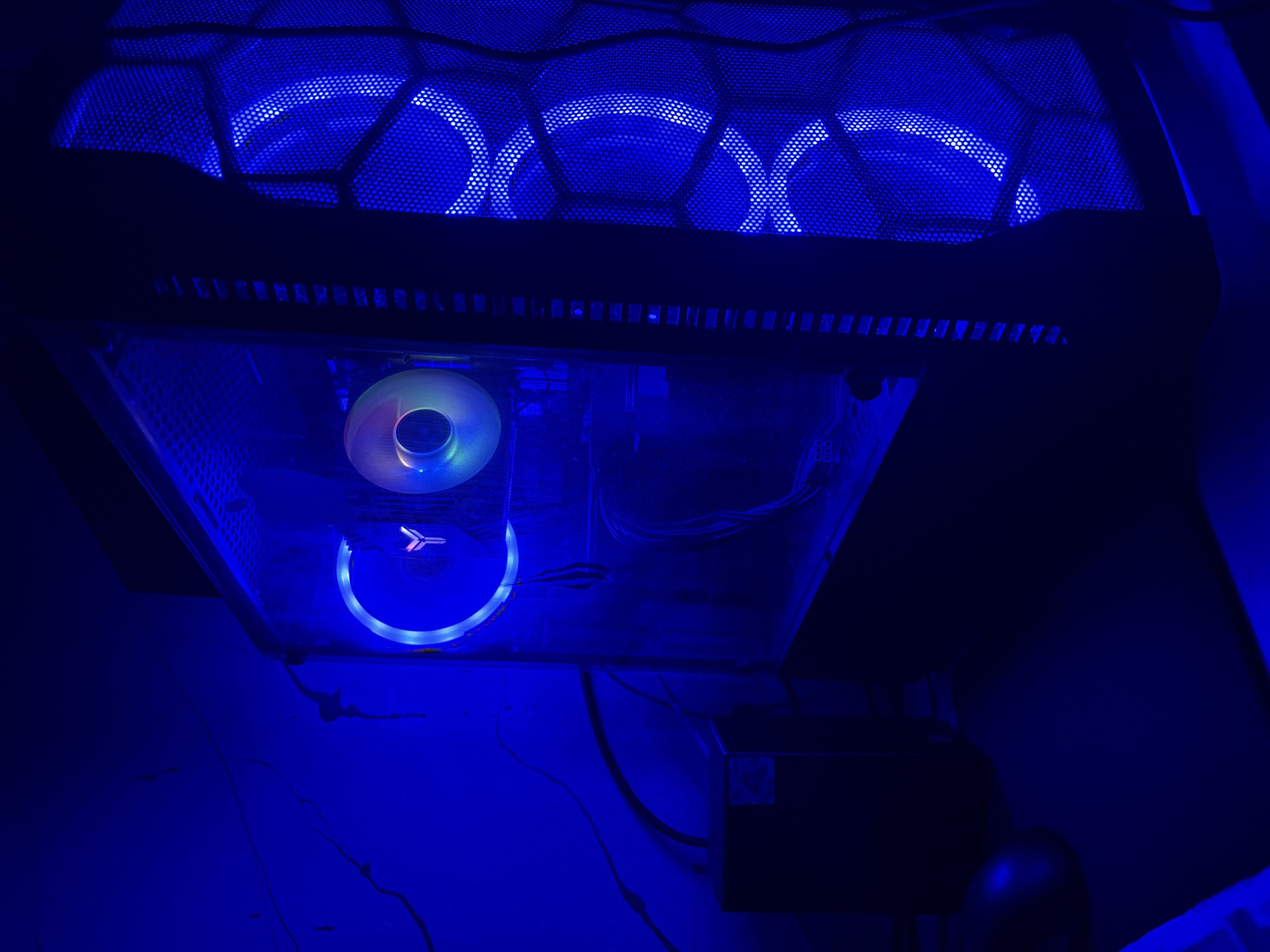 The image shows a gaming computer setup in a dimly lit room, with prominent blue LED lighting. The computer tower has a clear side panel, revealing internal components and a glowing fan with a 'Y' logo. The front of the tower also features illuminated hexagonal mesh. A black computer mouse is visible on a black mousepad in the foreground. The background wall is dark and appears to have some textured or painted elements. The lighting creates a strong blue hue throughout the scene. There are no people or animals present in the image.