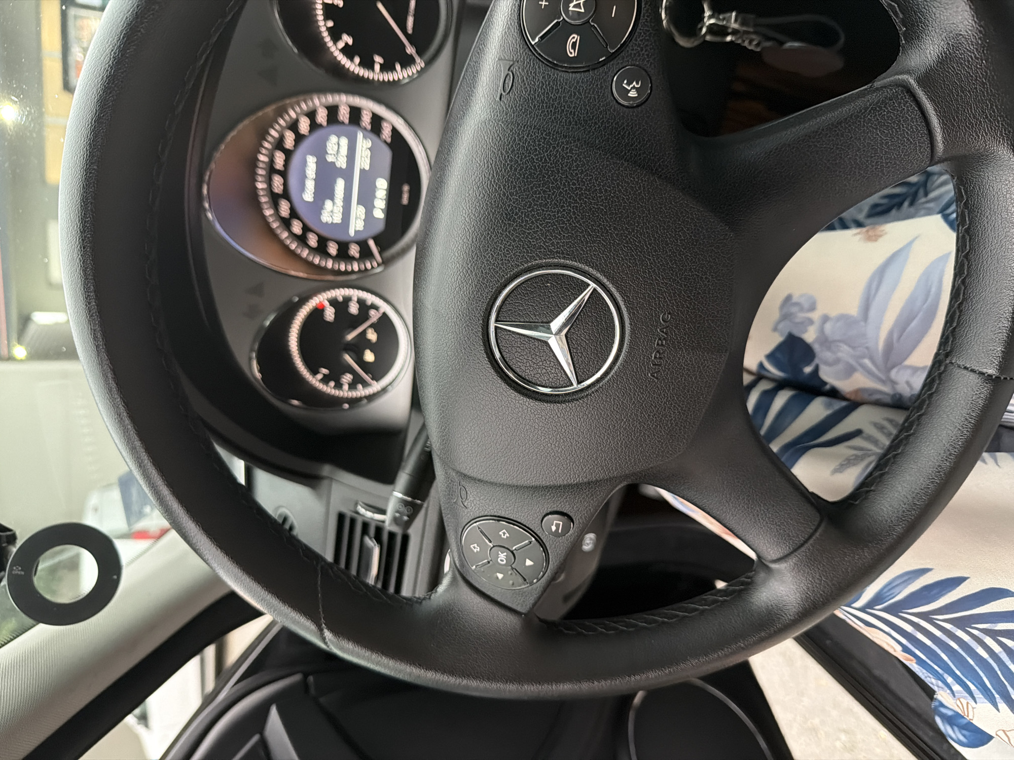 A close-up, high-angle shot captures the interior of a car, focusing on the steering wheel and dashboard from the driver's perspective. The black leather steering wheel, prominently featuring a silver Mercedes-Benz star logo in its center, takes up a significant portion of the frame. The word "AIRBAG" is visible on the right side of the steering wheel's hub.
The steering wheel features multi-function controls on both spokes. On the left spoke, a circular directional pad with "OK" in the middle, along with symbols for phone calls, volume, and navigation, is visible. Above this, individual buttons display a horn symbol and a music note. The texture of the plastic controls contrasts with the stitched leather of the steering wheel rim.
Behind the steering wheel, a partially visible instrument cluster includes three circular gauges. The central gauge is a digital display showing various trip computer readings: "From start", "1/20", "22.5 C" (temperature), "20 min", "31.9", "10.3 l/100km" (fuel consumption), and "00:00". Below the instrument cluster, air vents are visible.
Through the gaps in the steering wheel and to its right, a car seat or cushion upholstered in a patterned fabric with blue, white, and brown floral or leafy designs is visible. On the far left, a black circular ring, possibly a mount, is attached to the windshield or dashboard. The scene depicts a car that is likely stationary, with its systems powered on.