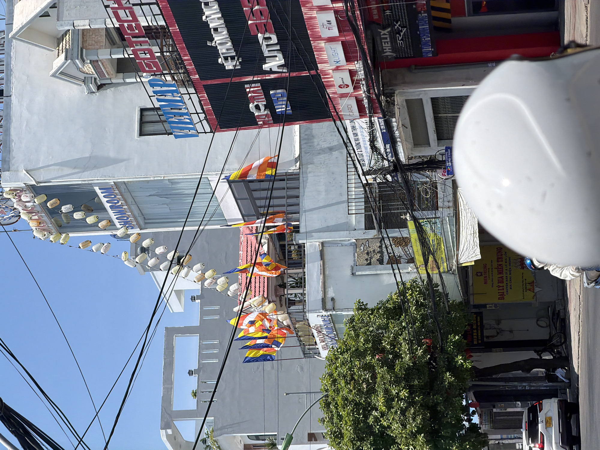 In Bac Nha Trang Ward, Vietnam, a street scene unfolds under a clear blue sky. A white SUV is parked on the left, facing away from the camera. To its right, a mature tree with dense green foliage stands tall, its branches laden with small red objects. In the foreground, the white helmet of a person on a motorbike is visible, obscuring their face and upper body.
Buildings line the street, characterized by white and light-colored facades. One building displays Buddhist prayer flags in yellow, orange, and white, fluttering in the breeze. Another building features a prominent "SHOWROOM" sign in blue letters. Higher up, a string of decorative lanterns hangs, alternating in color between white and orange. Further to the right, a building has bold red and black signage with white lettering that includes "VIETMAP" and "GOTECH". Other signs on this building appear to advertise car-related services, with brand names like "AUTO 365", "HELIX", and "TITAN".
A network of black electrical wires crisscrosses the sky above the buildings, creating a tangled canopy. The street itself appears to be paved. The overall impression is a bustling urban environment with a mix of commercial establishments and religious symbols, typical of a Vietnamese city.