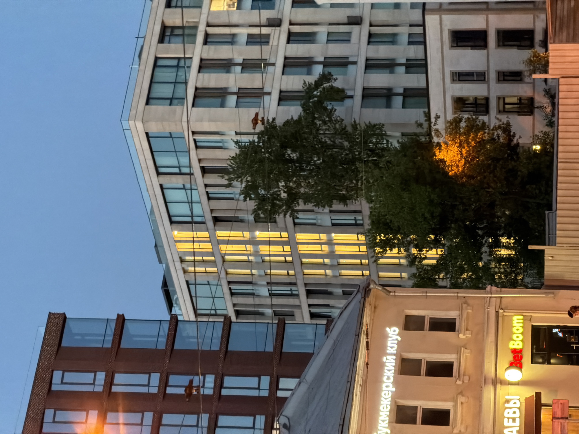 The image depicts a street scene in Moscow, Russia, at dusk. Three buildings of varying heights and architectural styles dominate the frame. The tallest building has a modern design with large windows, some of which are illuminated. A tree partially obscures this building. Another building has a brown, brick-like facade with reflective glass. A third building has a beige facade and features the words "букмекерский клуб" (bookmaker club) vertically, along with the word "Boom." There is evidence of exterior construction or maintenance in the form of a worker visible on the side of one of the buildings. The sky is a light blue color, suggesting early evening.