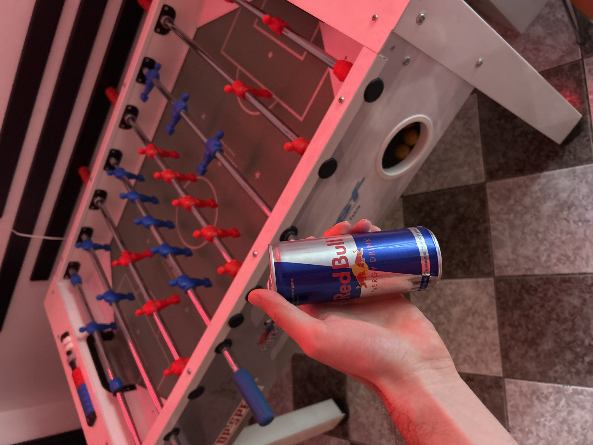 A close-up shot shows a hand holding a Red Bull can in front of a foosball table. The table is primarily white with red and blue players. Yellow foosballs are visible in a holding area. The background features striped patterns on the wall and tiled flooring. The image is likely taken indoors, and based on the Red Bull can, the setting may be a recreational area or a game room.