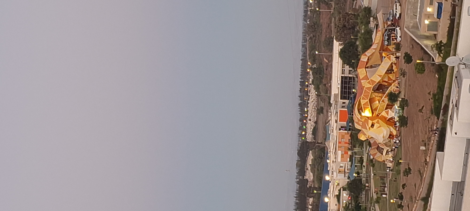This aerial view captures an exterior scene in Ashkelon, Israel, likely during twilight or early evening, as evidenced by the soft, diffused light and some illuminated structures. The focal point of the image is a large, abstract, geometric structure that resembles a stylized lion. This structure, predominantly gold and orange, is a prominent feature in what appears to be a public space or park.

Scattered around the lion structure are numerous small figures, presumably people, gathered in groups. The area around the structure is landscaped with low-lying bushes and trees. In the background, there are several modern buildings with white and orange facades, suggesting a developed urban or suburban area. Some of these buildings have illuminated windows.

Further in the distance, a line of trees forms a dark silhouette against the sky, and beyond that, a hazy horizon with a faint, tall structure, possibly a tower or antenna, is visible. The sky itself is a soft gradient of pale grey to light blue, indicating the transition from day to night. Several streetlights with yellow lights are also present in the scene, illuminating pathways and open areas. Some vehicles, including cars and a van, are parked near the buildings in the background. The overall impression is of a modern development within the city of Ashkelon, with the lion structure serving as a distinctive landmark.