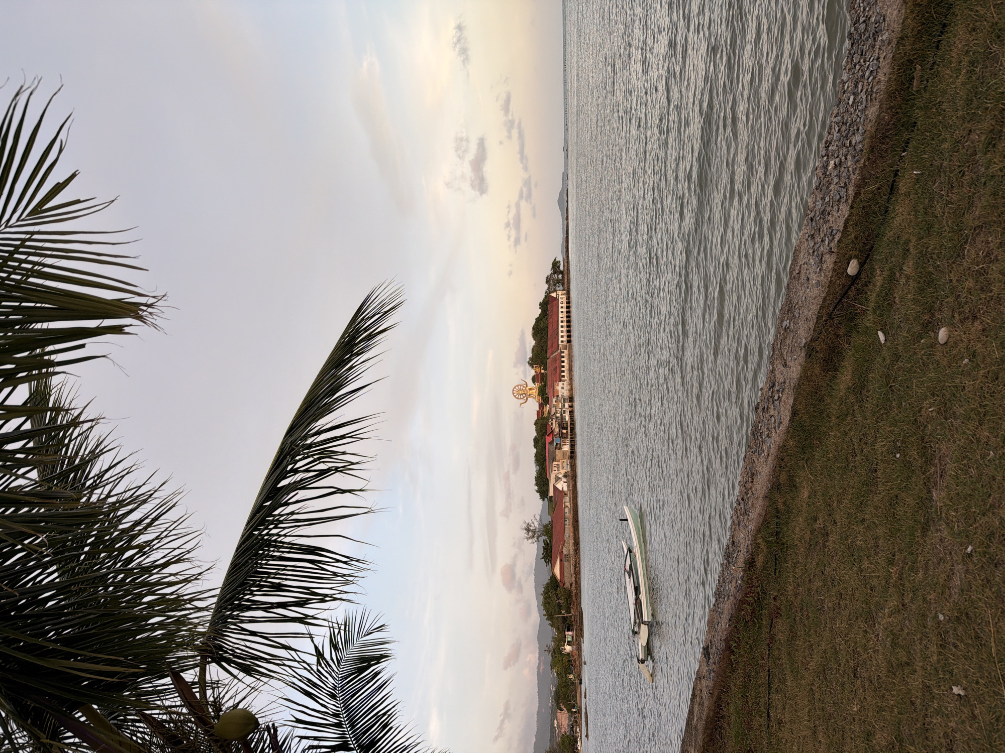 An outdoor, eye-level, vertical medium shot captures a tranquil coastal scene in the city of Ko Samui District, Thailand, during what appears to be late afternoon or early evening. The foreground is dominated by a patch of dry, brown grass meeting a concrete and stone embankment. Beyond the embankment, the calm, grey sea stretches towards the horizon, its surface gently rippled. A small, light-colored boat, possibly a catamaran, floats in the water in the mid-left portion of the frame, appearing unoccupied.
In the background, a coastline is visible, lined with buildings featuring red-tiled roofs, nestled amongst lush green trees and hills. A prominent, large golden statue of a religious figure, possibly Buddha, stands tall on a hill, overlooking the town. The sky above is a soft gradient of pale blue and grey, with scattered, wispy clouds tinged with a hint of pink and orange from the setting sun. A portion of a palm tree, with its long, dark green fronds, frames the top left of the image, adding a tropical element to the serene vista. There are no people or animals clearly visible in the scene.