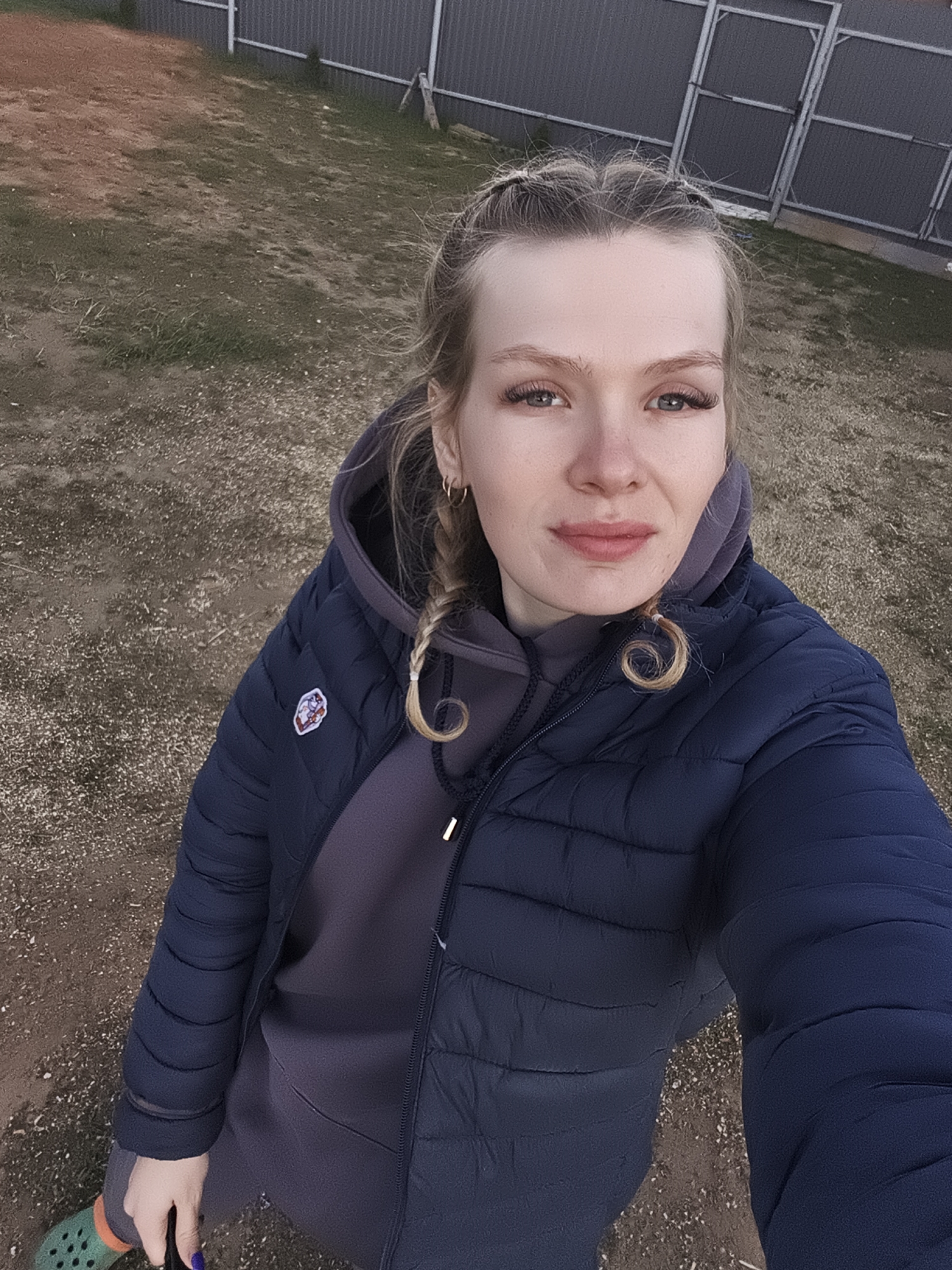 The image is a selfie of a young woman with blonde hair styled in braids, wearing a navy blue puffer jacket over a gray hoodie and sweatshirt. She is standing outdoors on a patch of ground that appears to be a mix of dirt, gravel, and sparse grass. In the background is a corrugated metal fence, possibly indicating a residential or industrial area. The photograph was taken in Русский Вожой, Russia.