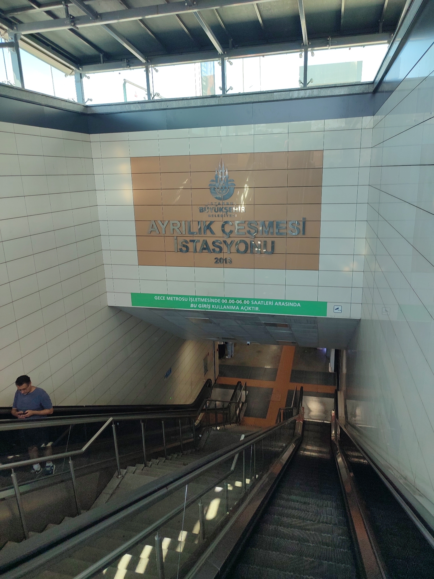 This is a medium-angle shot of an underground station entrance in Kadıköy, Turkey. The station is identified by a sign that reads "AYRILIK ÇEŞMESİ İSTASYONU" with the Istanbul Metropolitan Municipality logo above it, and the year "2013" below. A green banner with Turkish text indicates that the station is open for use between 00:00 and 06:00 for night metro operations.
In the foreground, a man wearing a blue t-shirt and glasses is standing on an escalator, looking down at his phone. He is positioned to the left of the frame. The escalators and staircases descend into the lower levels of the station. The walls of the station are tiled in a pattern of white and light brown squares. Above the tiled walls, there is a modern metal and glass roof structure, allowing natural light to enter the station. The floor of the lower level features an orange crosswalk marking.