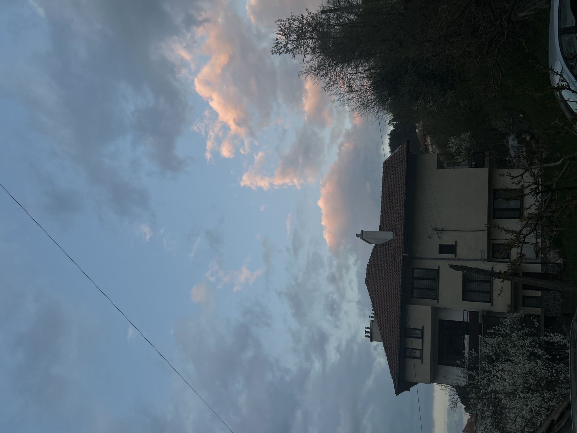 The image presents an exterior view, likely taken at dusk. A two-story building with a red-tiled roof is positioned on the right side of the frame. The building's facade is light-colored with dark-framed windows. A black balcony is visible on the first floor. To the left of the building, the sky is filled with grey and pink-tinged clouds. The upper portion of the image is partly obscured by trees with bare branches. A wire runs diagonally across the lower left part of the sky. The location is potentially Zlatograd, Bulgaria, but this cannot be conclusively confirmed without further information. No people are visible.